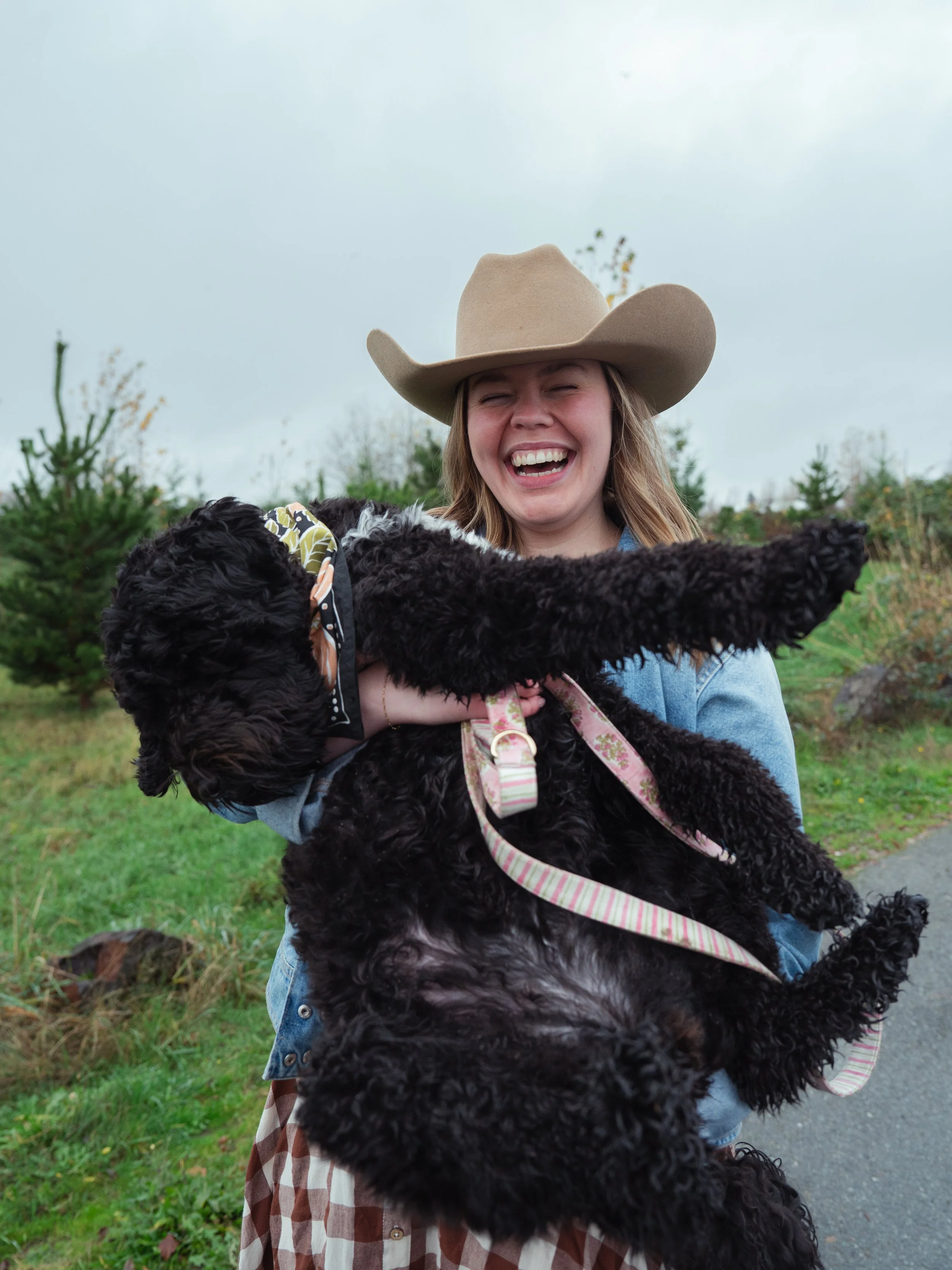 A woman wearing a cowboy hat and a denim jacket holding a black curly-haired dog with a pink and white leash outdoors on a cloudy day.