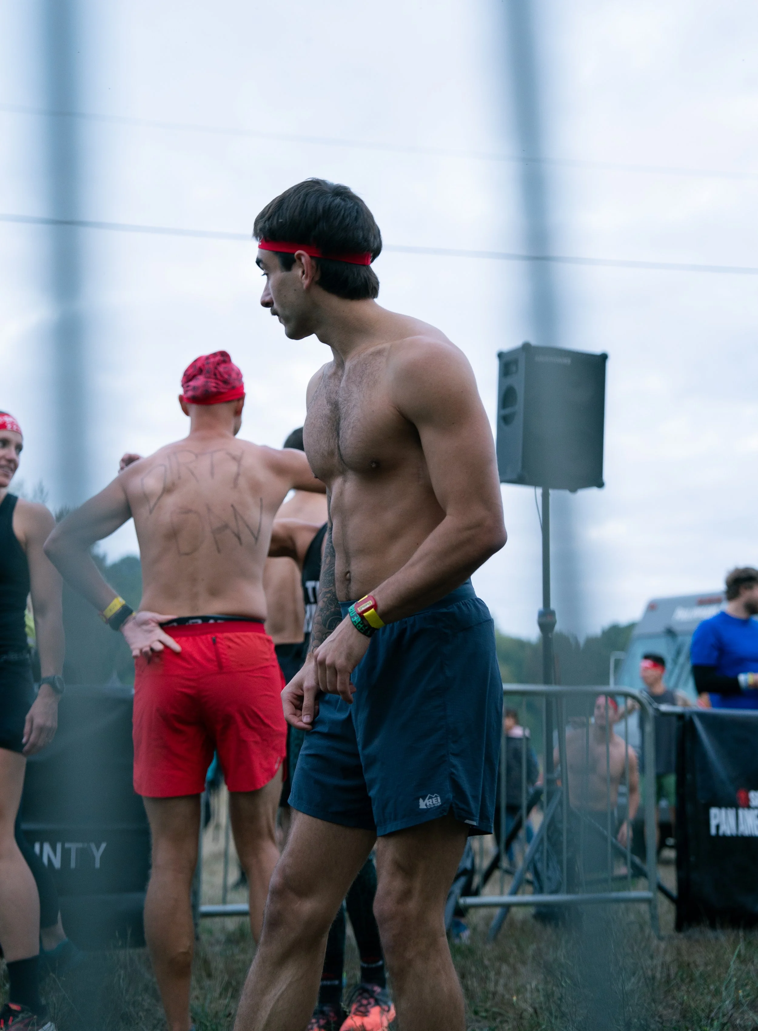 A group of shirtless male runners at an outdoor event, some wearing red headbands and others with messages written on their backs, preparing for or after a race.