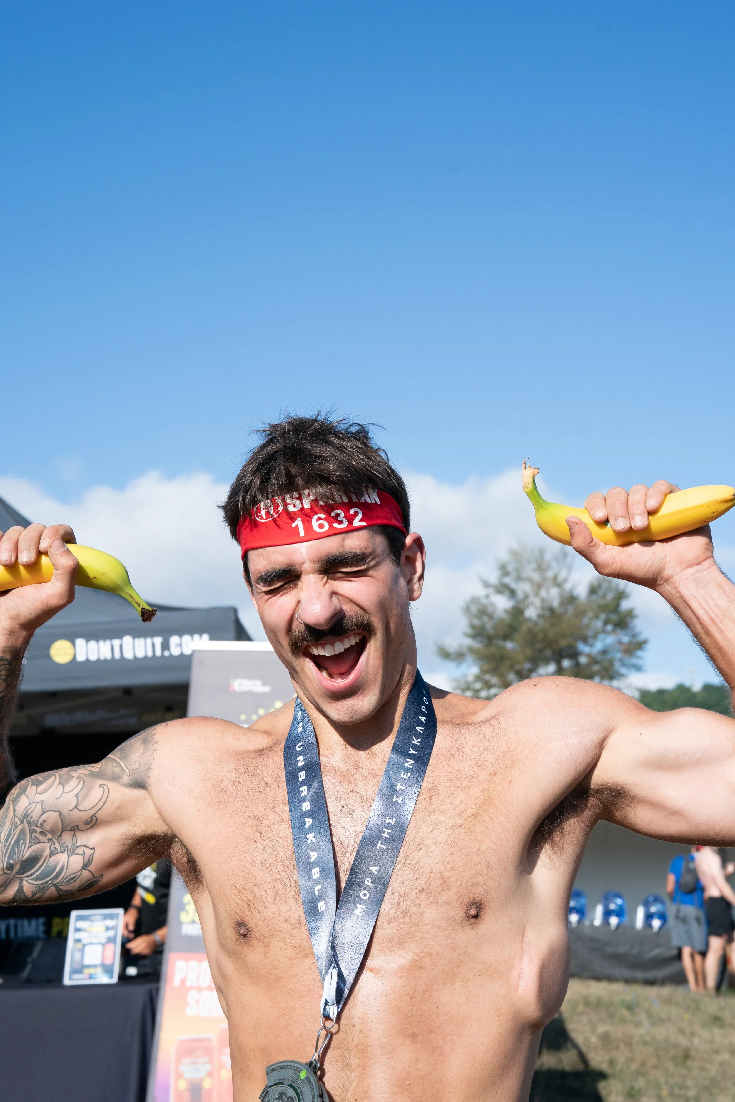 A shirtless man with a mustache celebrating after a race, holding bananas in both hands, wearing a red headband, and a medal around his neck, outdoors on a sunny day.