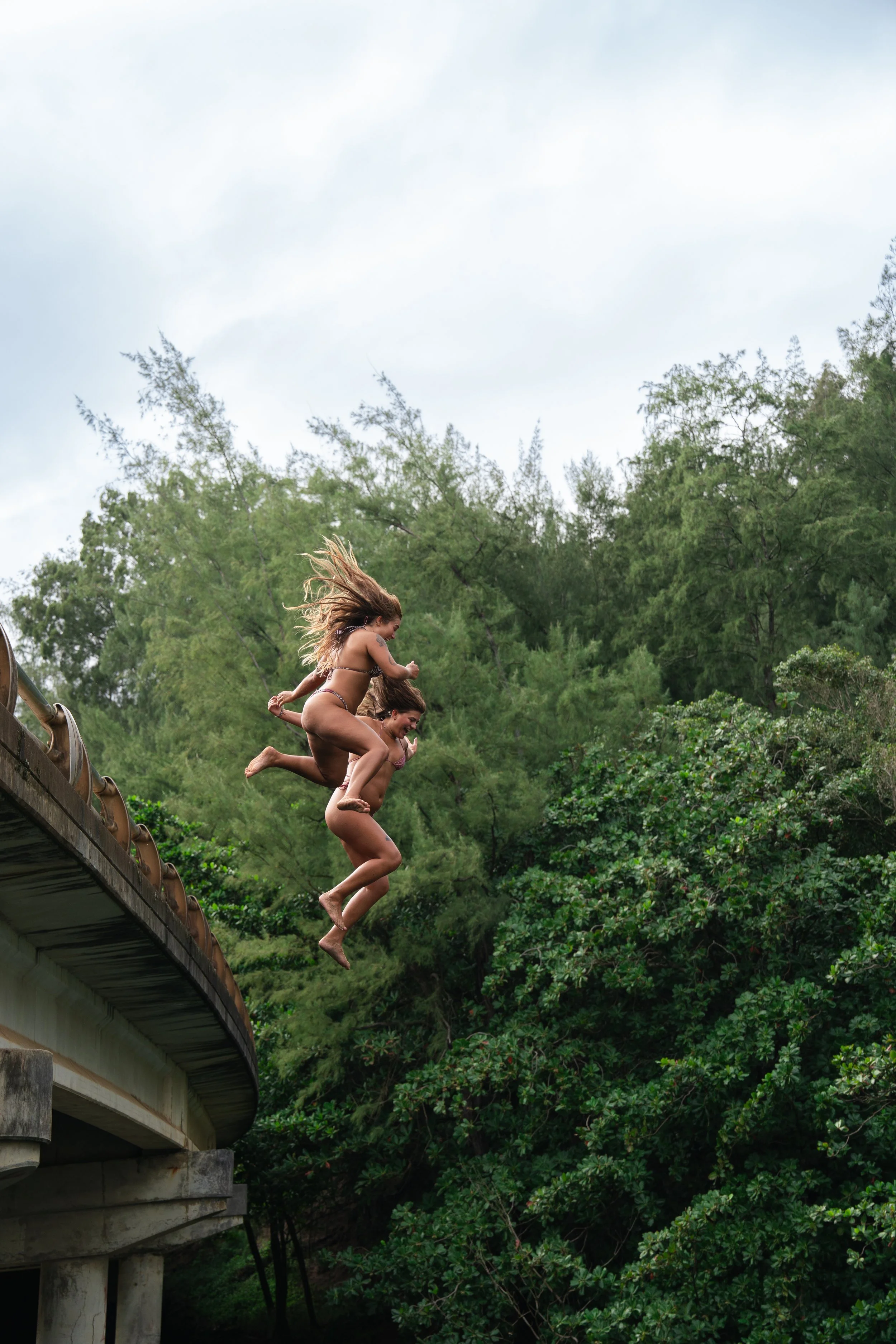 Two women jumping off a bridge into a river surrounded by green trees.