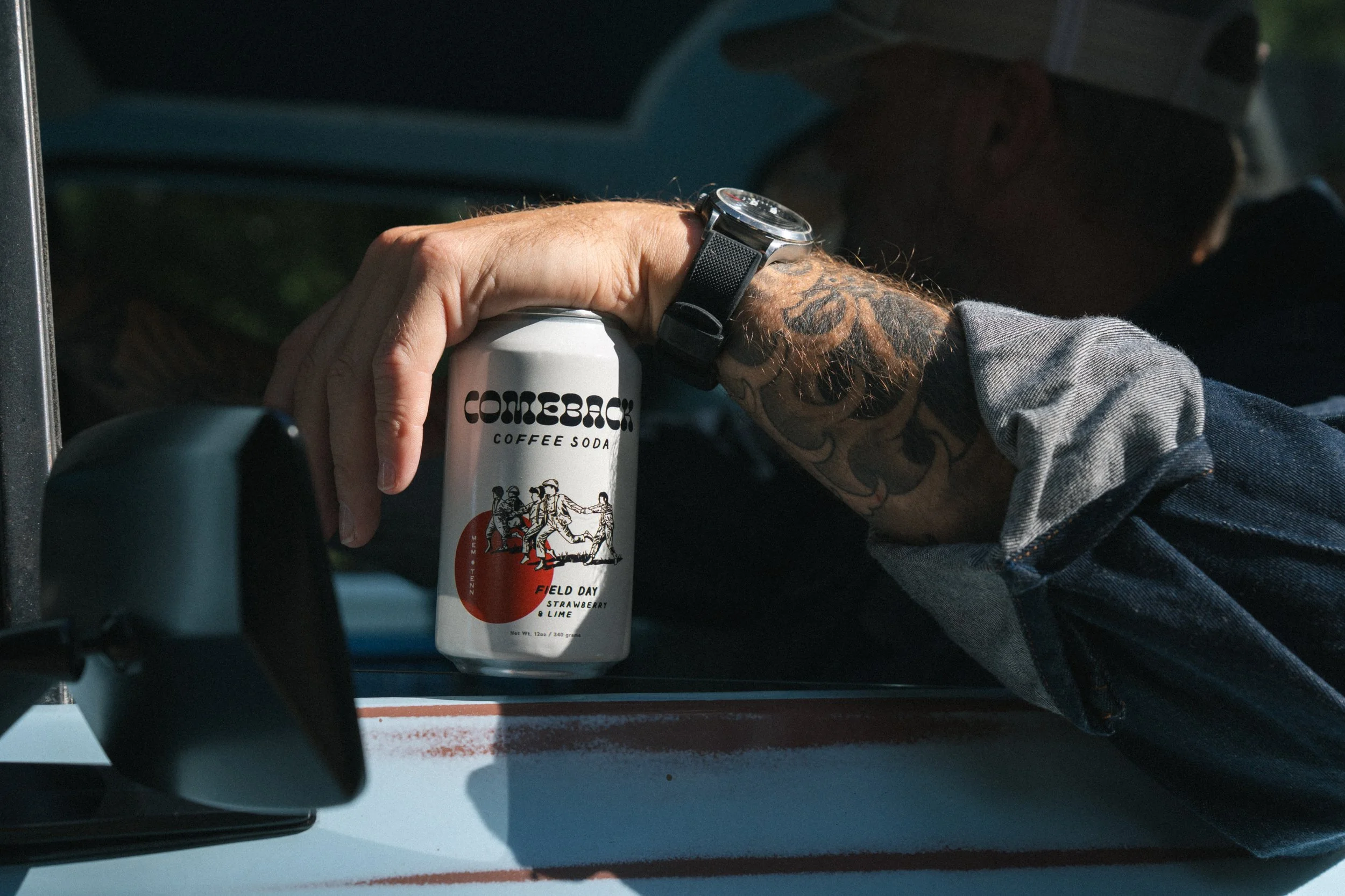 A person's hand with tattoos holding a can of 'Comeback Coffee Soda' while leaning out of a vehicle window.