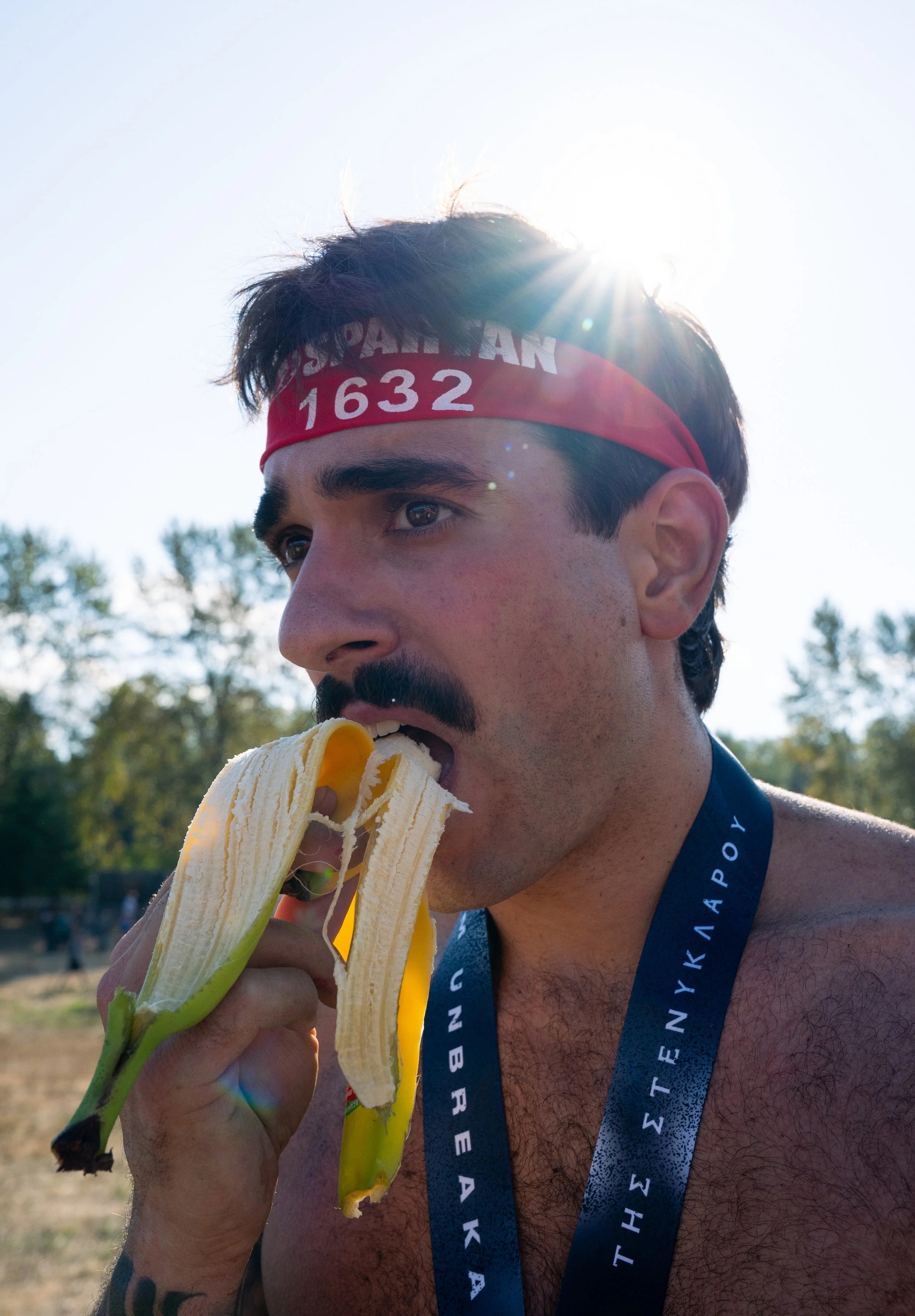A man with a mustache and dark hair, wearing a red headband with the text 'SPARTAN 1632', eats a peeled banana outdoors, with a medal around his neck and trees in the background, sunlight shining behind his head.