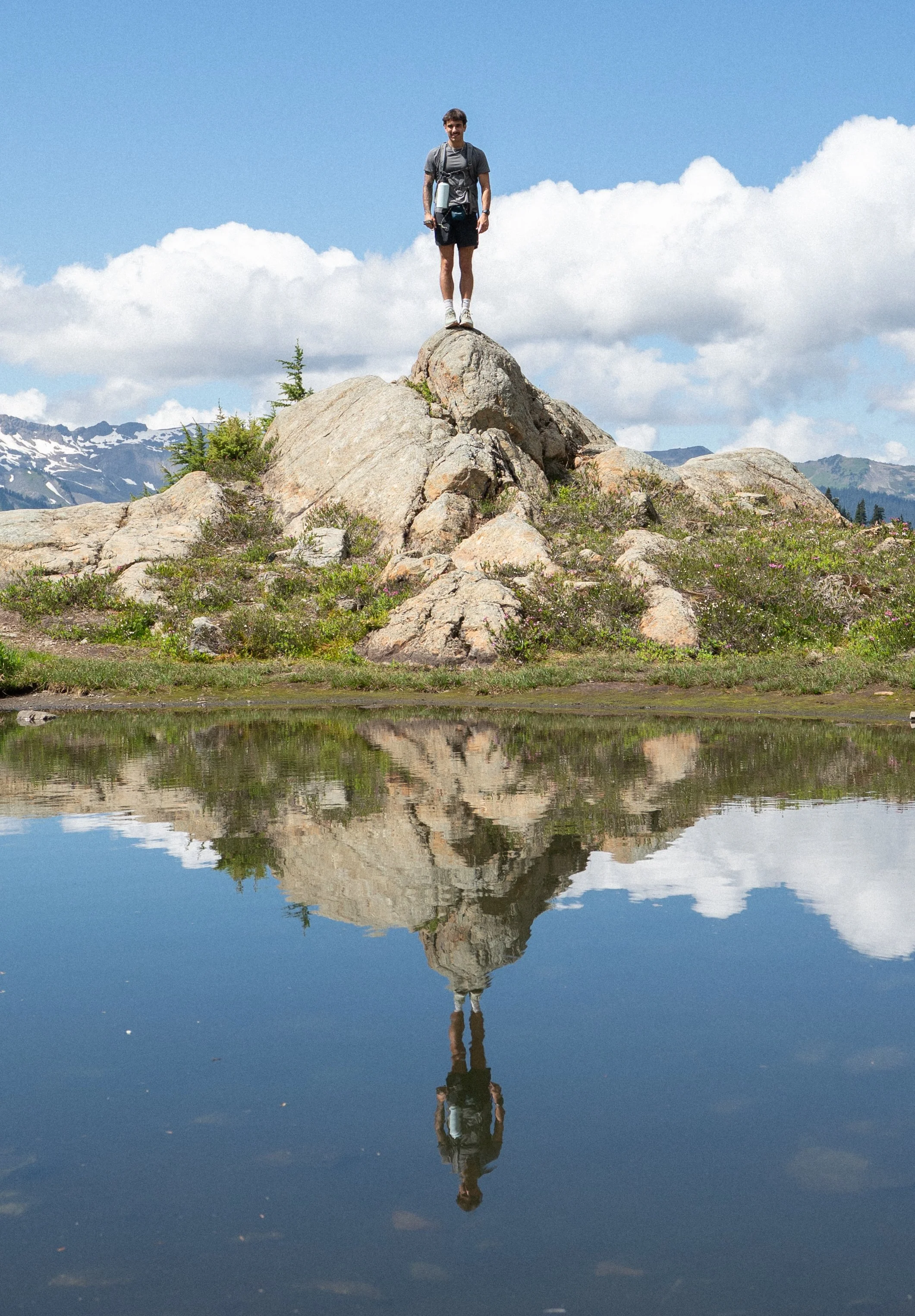 A man standing on a large rock formation near a small lake, with mountains and a blue sky with clouds in the background.