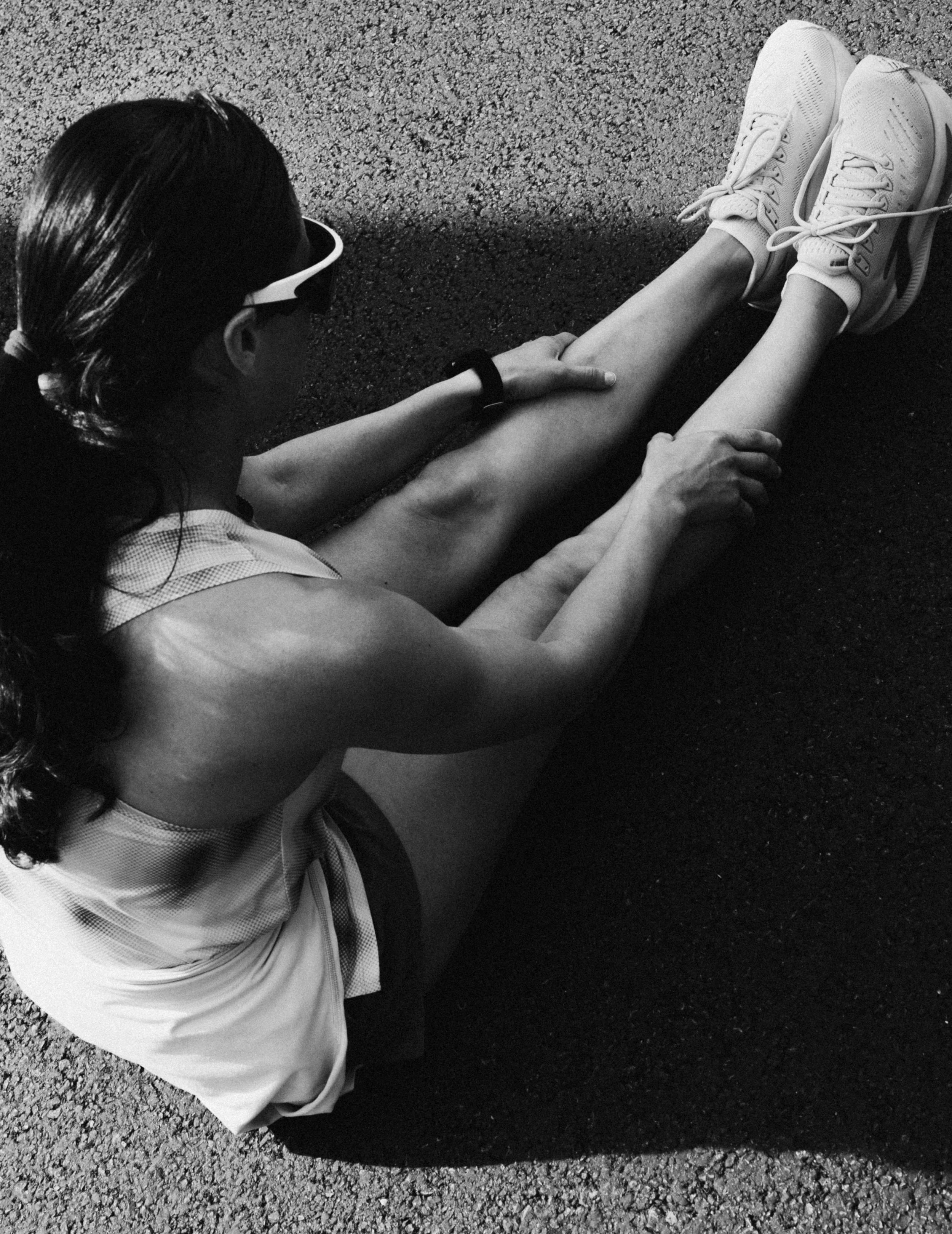 A woman wearing athletic clothing and running shoes, sitting on the ground stretching her legs, with sunlight casting shadows on the textured surface.