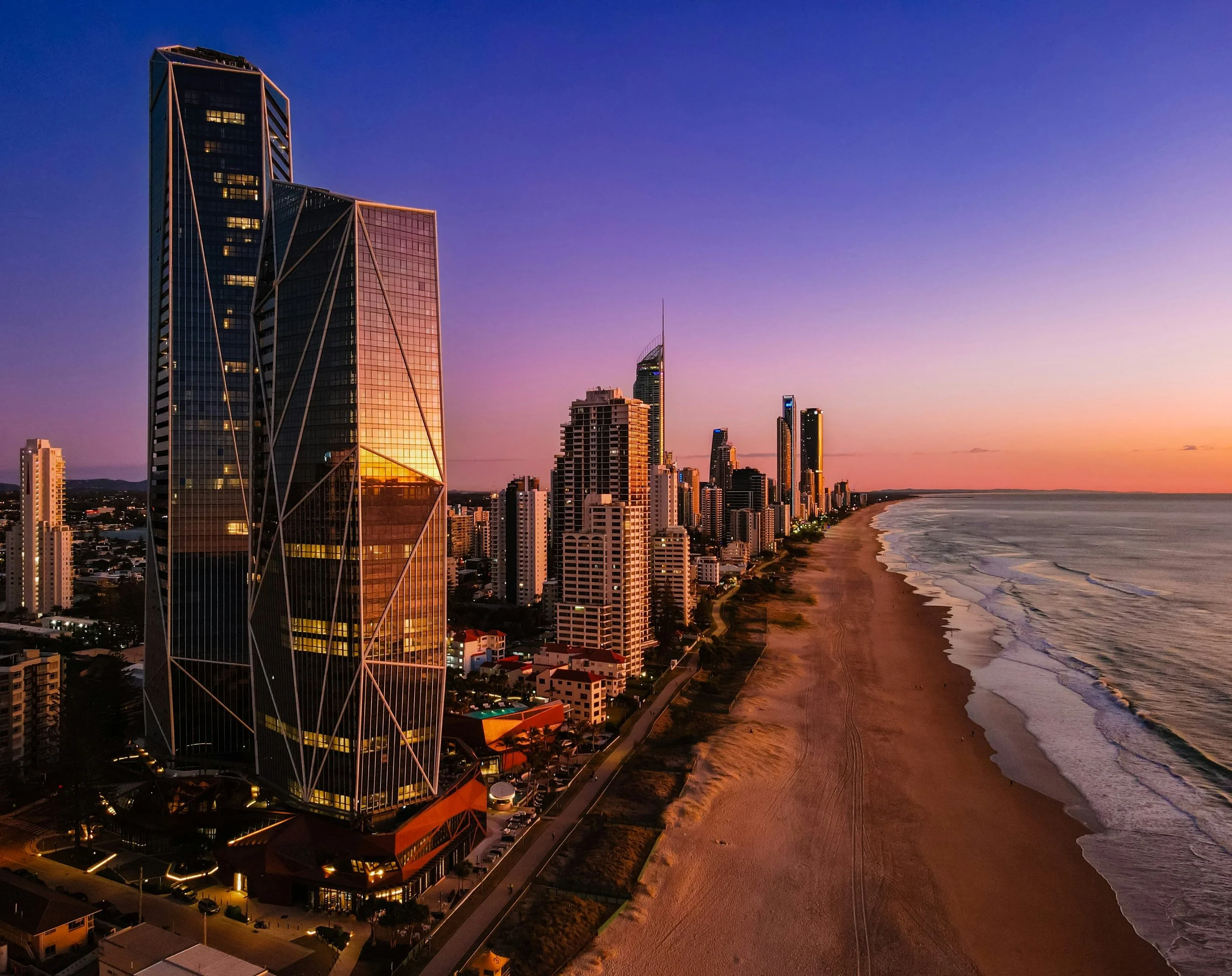 A city skyline with tall skyscrapers along a shoreline at sunset, reflecting orange and purple hues in the sky.