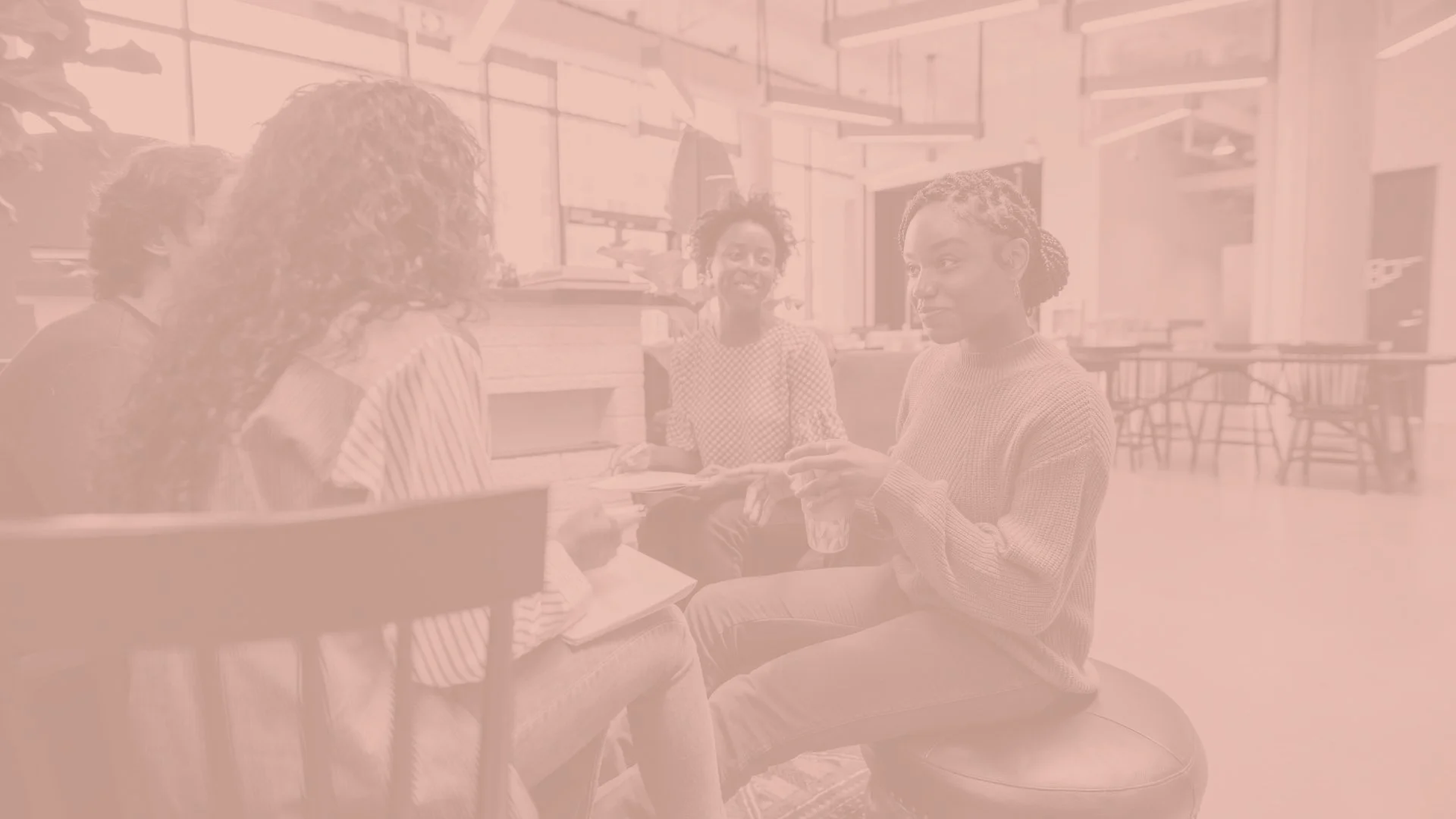 Four women sitting together in a bright indoor space, having a conversation, some holding notebooks and glasses, smiling and engaged.