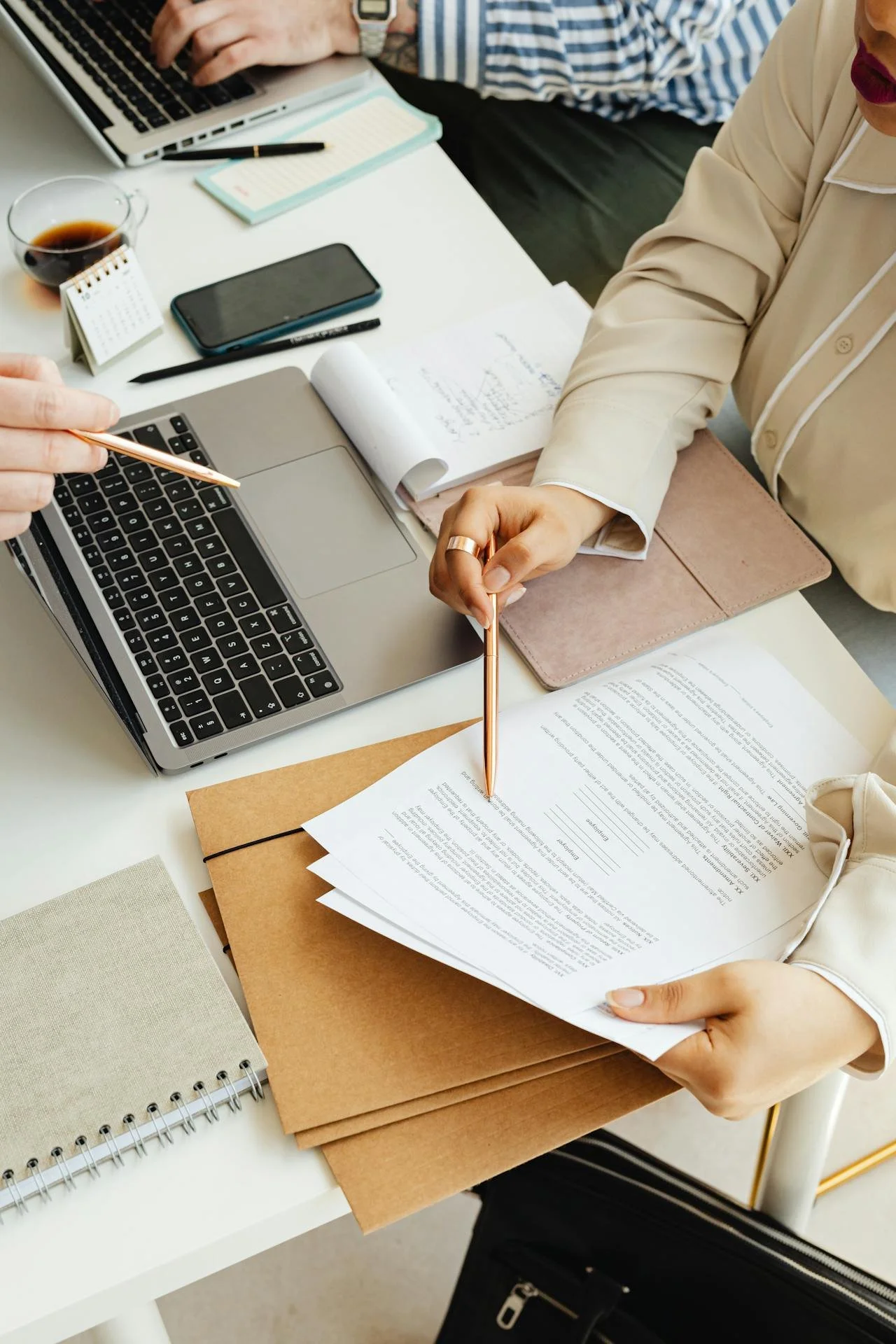 People working at a desk with laptops, notebooks, documents, a phone, and a glass of coffee.