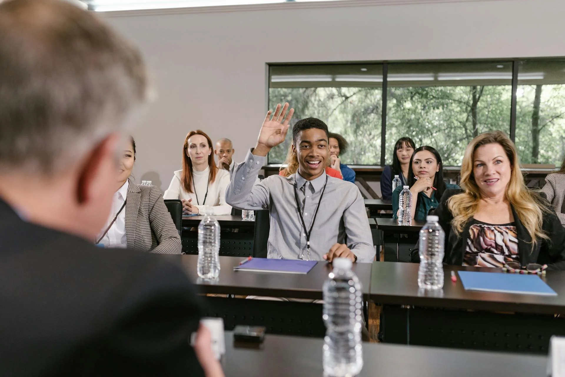 A young man with short hair raising his hand during a conference or classroom session. He is smiling and sitting at a table with water bottles and notebooks, with other diverse attendees listening attentively in a modern room with large windows and greenery outside.