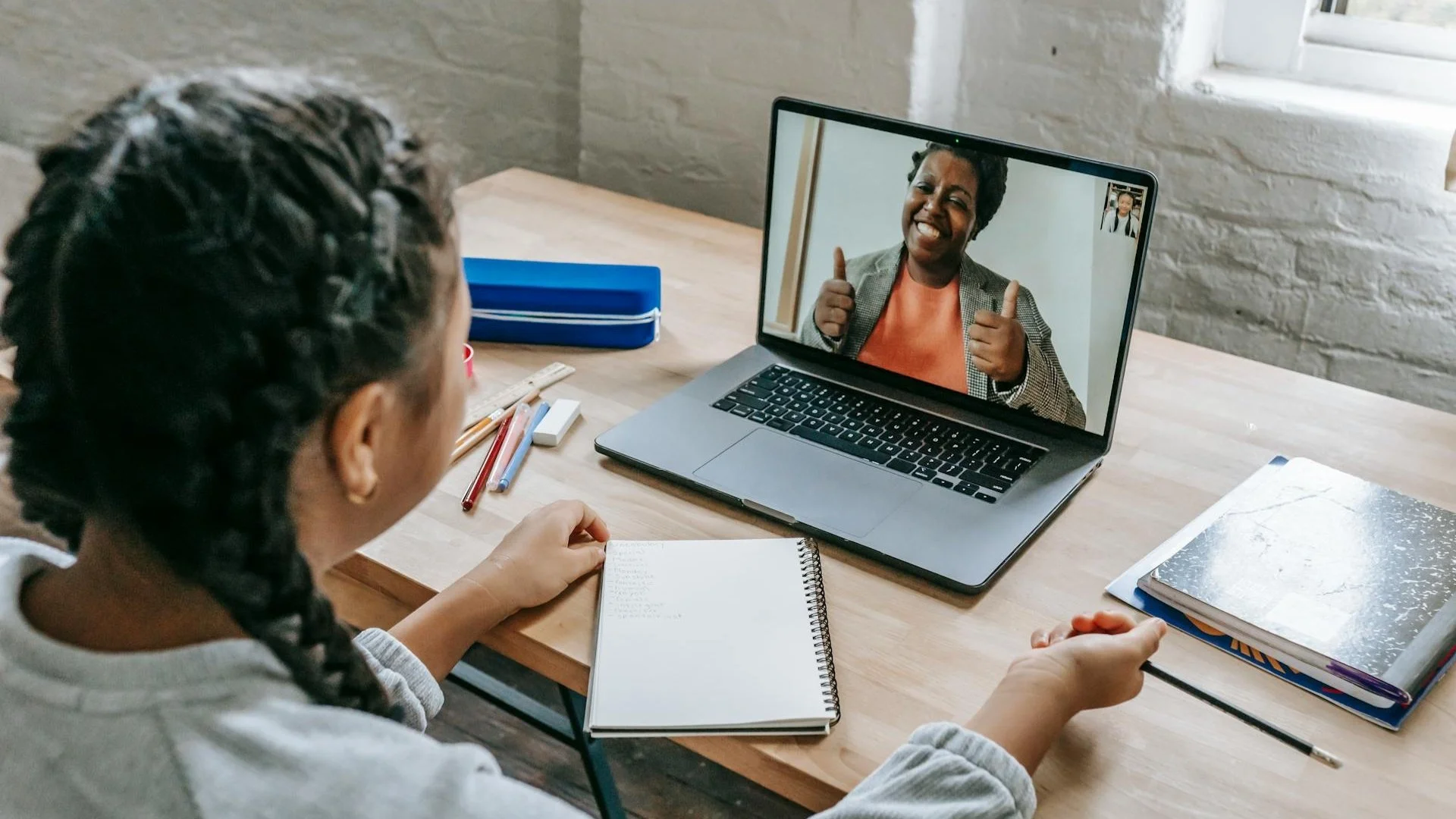 A girl with braided hair participating in a video call on her laptop with a woman giving a thumbs up and smiling.