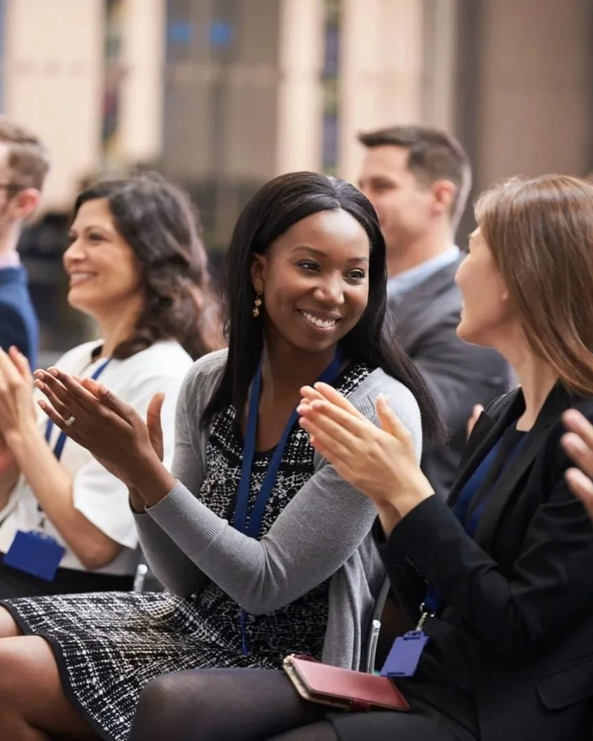 People at a professional conference or seminar, engaged in conversation and clapping, wearing business attire and conference badges.