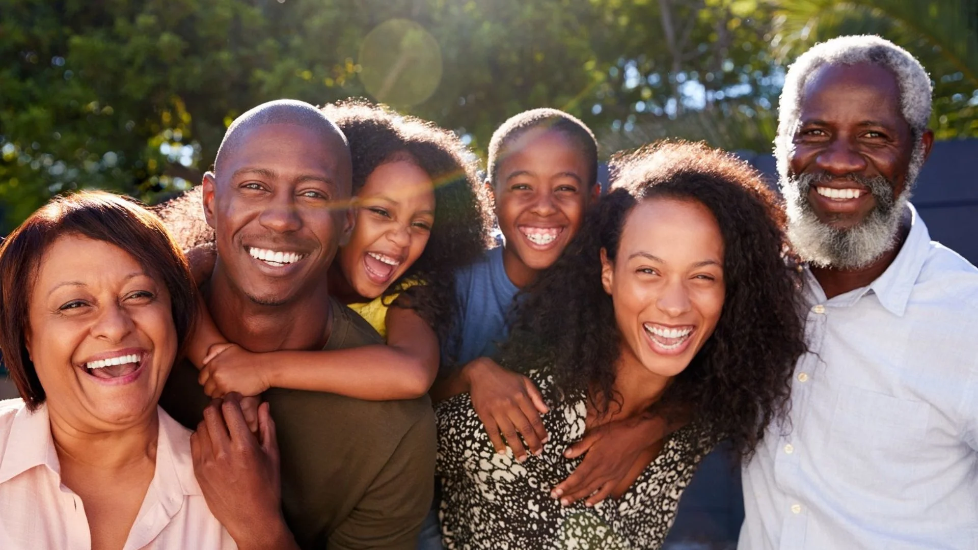 Family of six people smiling and laughing outdoors with trees and sunlight in the background.