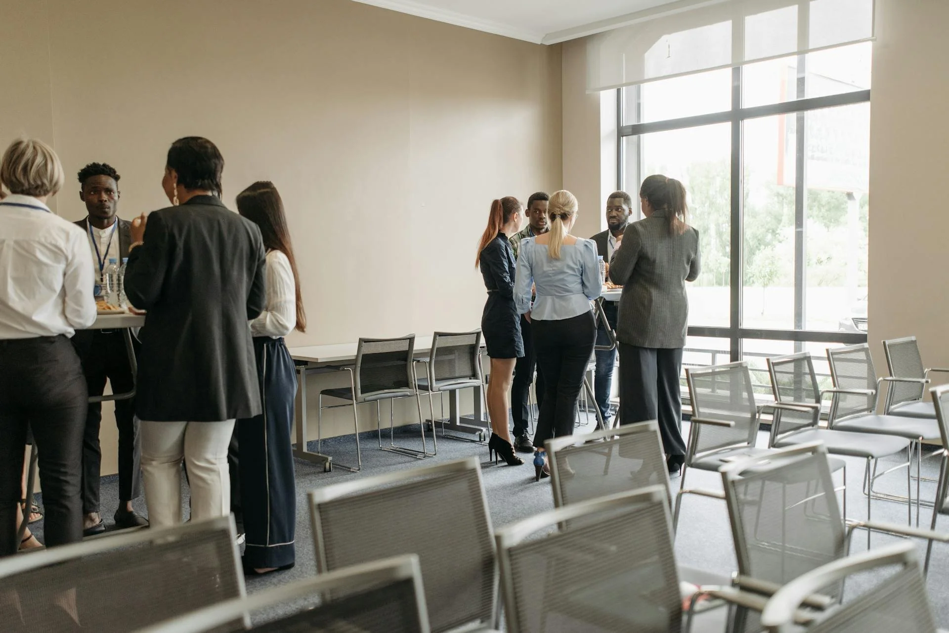 Business professionals gathered in a conference room, some standing and talking near a window, others around a high-top table, with empty chairs and large windows in the background.