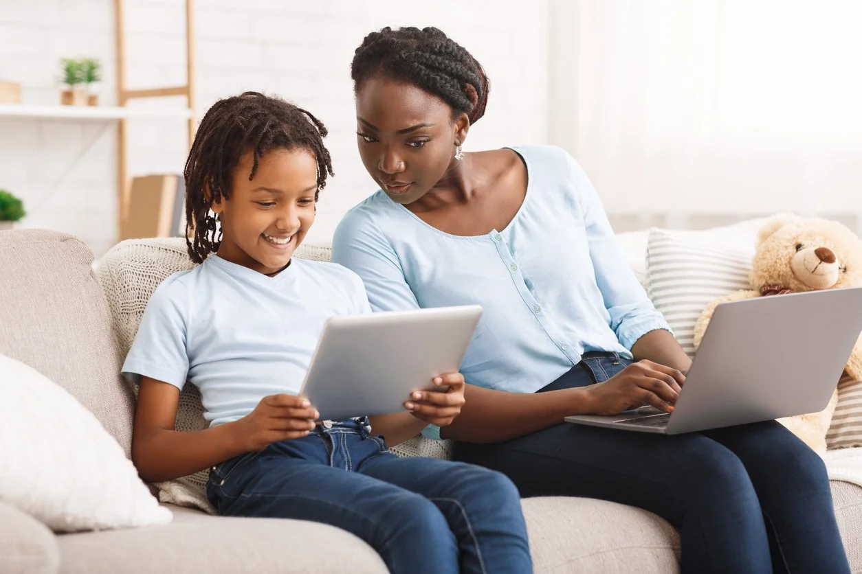 A woman and girl sitting on a sofa, looking at a tablet together, with a laptop on the woman's lap, in a bright living room with a teddy bear and pillows.