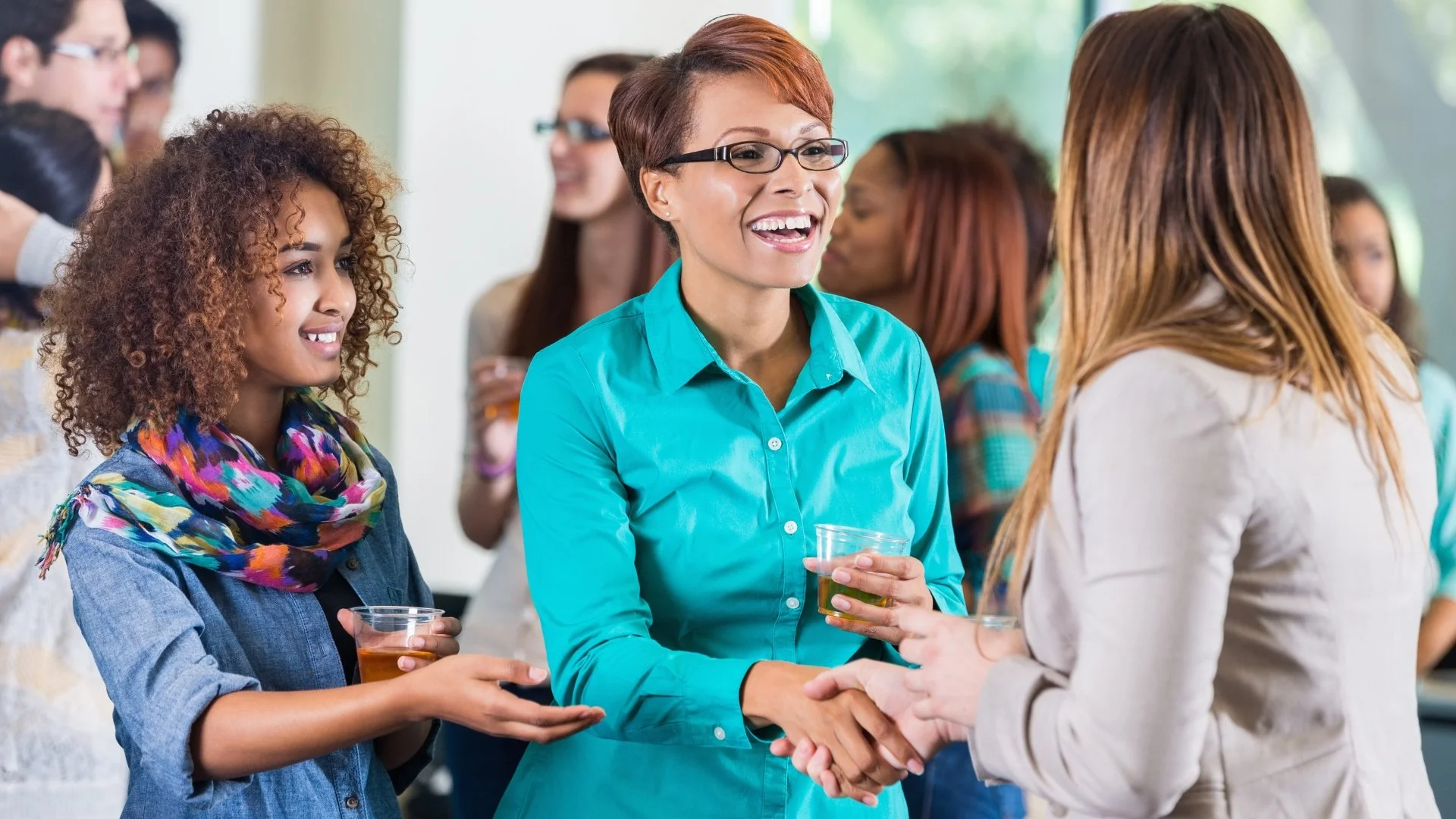 A group of diverse women at a social gathering, with one woman in a turquoise shirt smiling and shaking hands with another woman, while a third woman with curly hair and a colorful scarf watches.