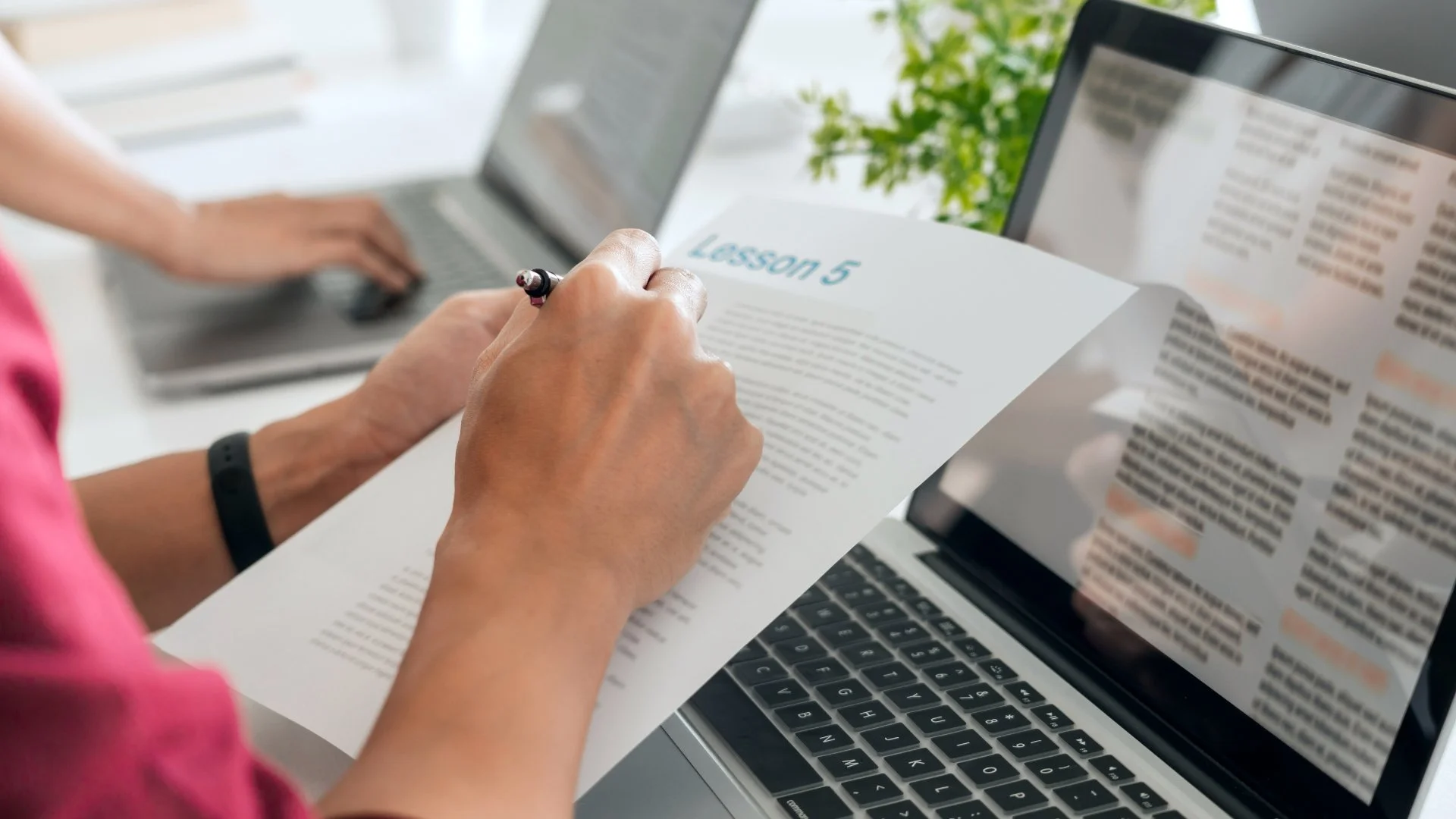 Person holding a paper titled 'Lesson 5' with one hand and writing with a pen, sitting at a laptop on a desk with another person using a laptop in the background and a green plant nearby.
