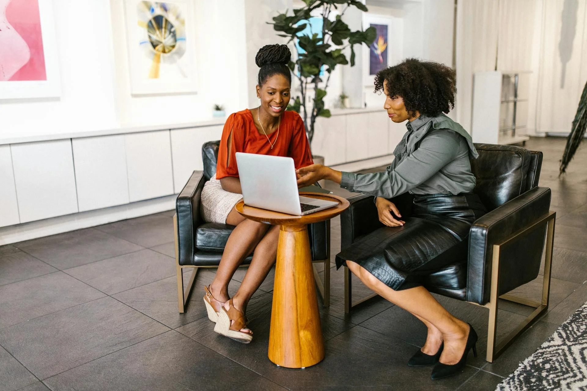 Two women sitting at a small round table with a laptop, engaging in a conversation in a modern art gallery or office lounge.
