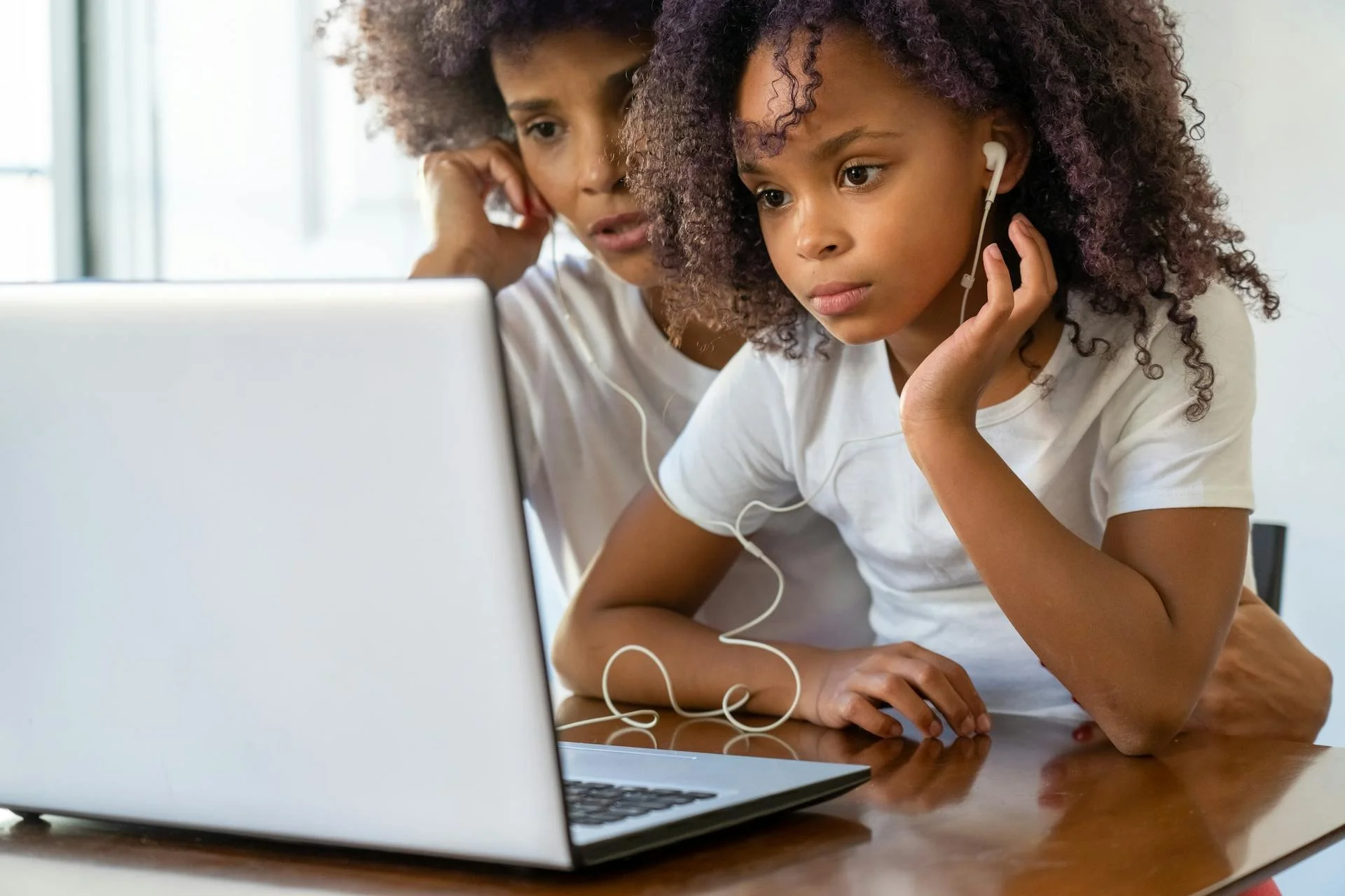 A young girl and an adult woman are looking at a laptop screen with worried expressions. The girl is wearing headphones and leaning forward, while the woman supports her chin with her hand.