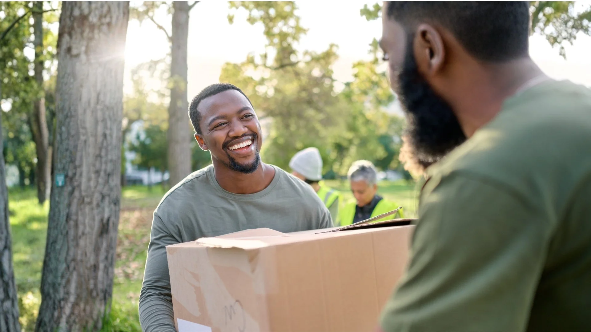 Two men exchanging a cardboard box outdoors in a park, smiling and engaging in conversation with sunlight filtering through trees in the background.