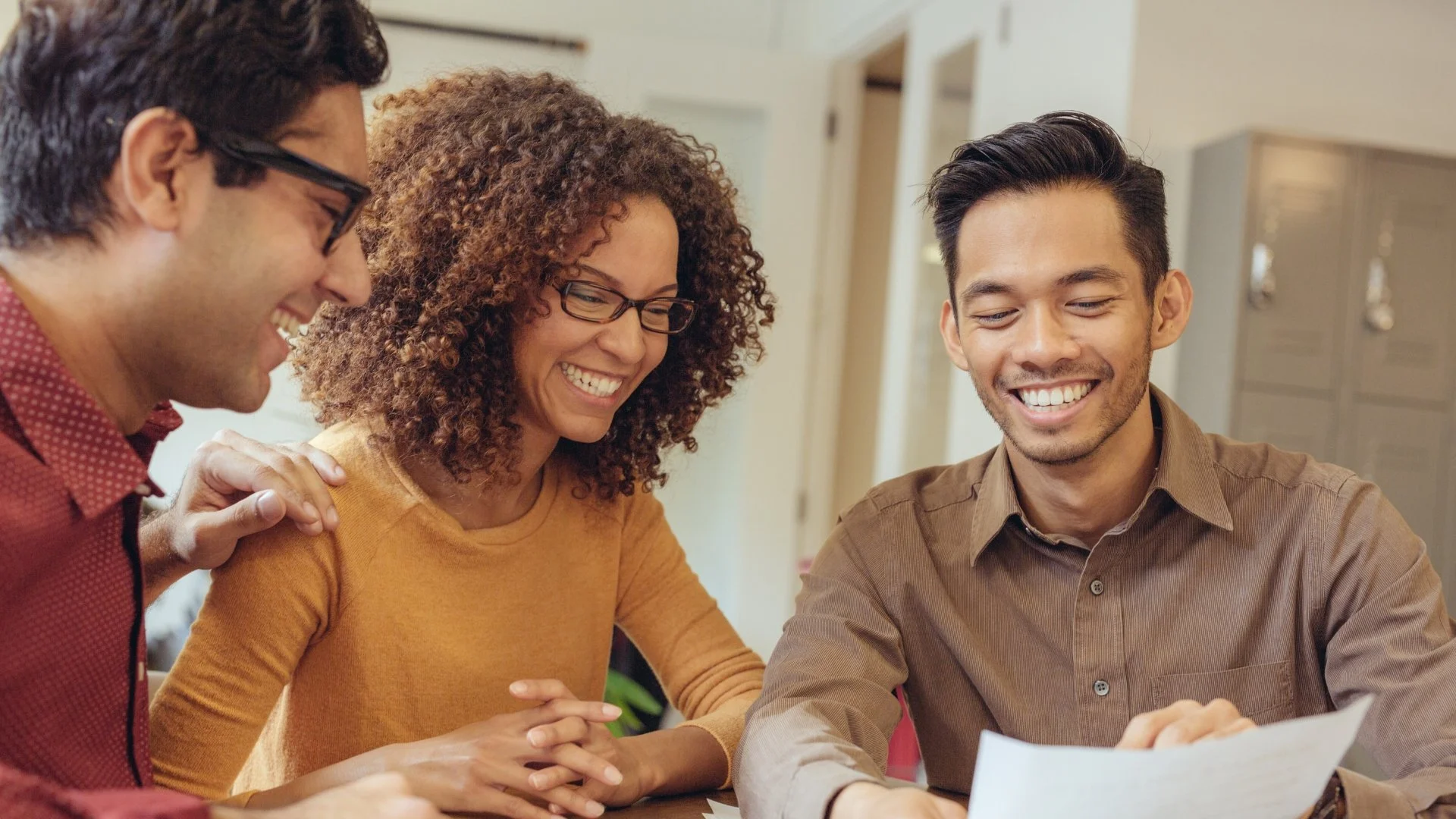 Three individuals smiling and looking at paper together in an office setting.