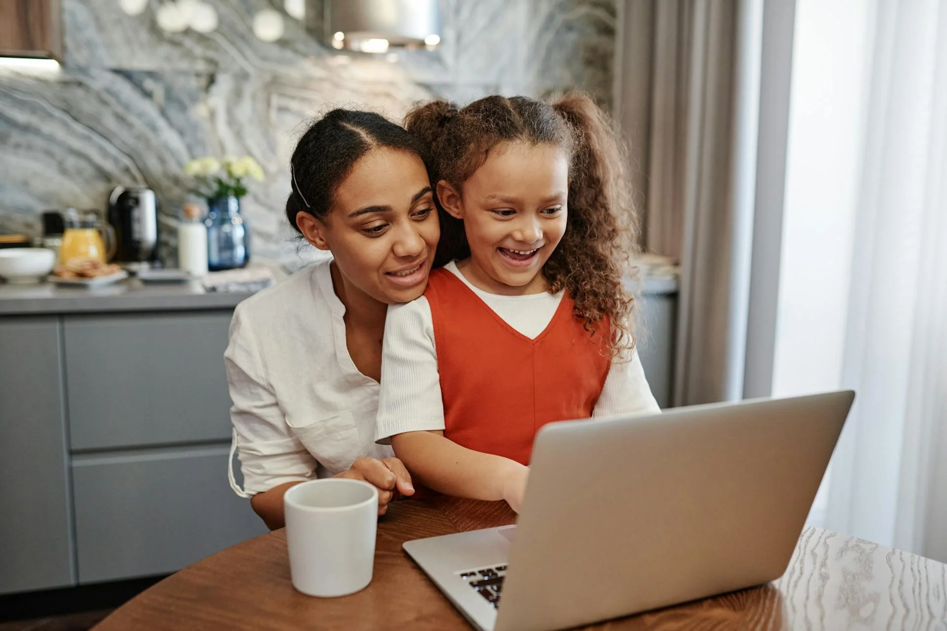 A woman and a young girl sit together at a table, looking at a laptop screen and smiling in a kitchen with a stone backsplash and modern decor.