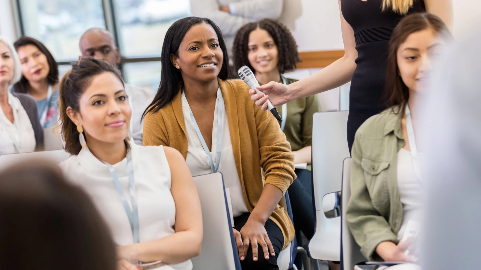 Woman in a mustard cardigan asks a question during a conference or workshop, with others listening, some taking notes, in a room with large windows.