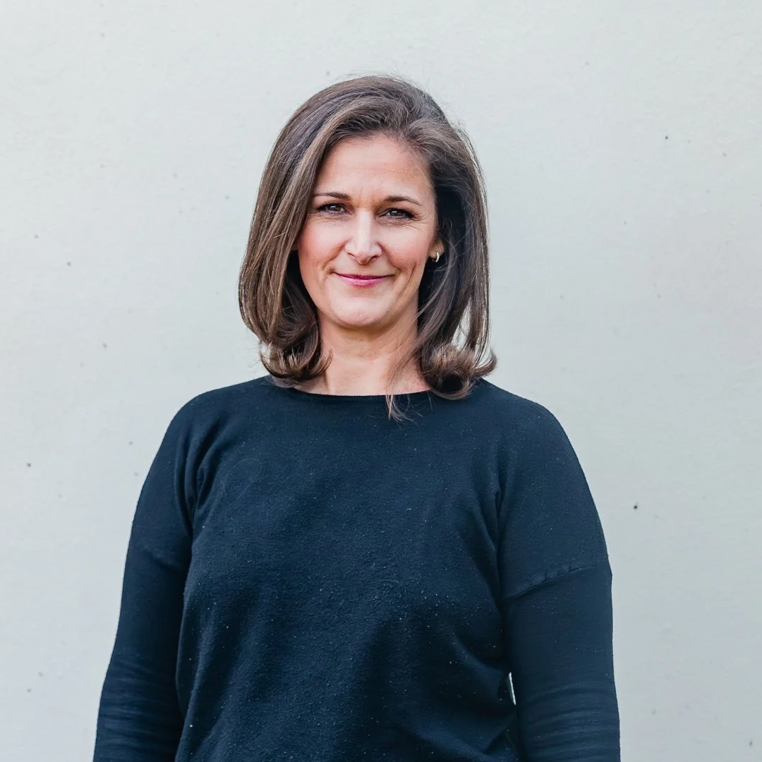 A woman with shoulder-length brown hair wearing a black long-sleeve shirt, standing against a light-colored wall.