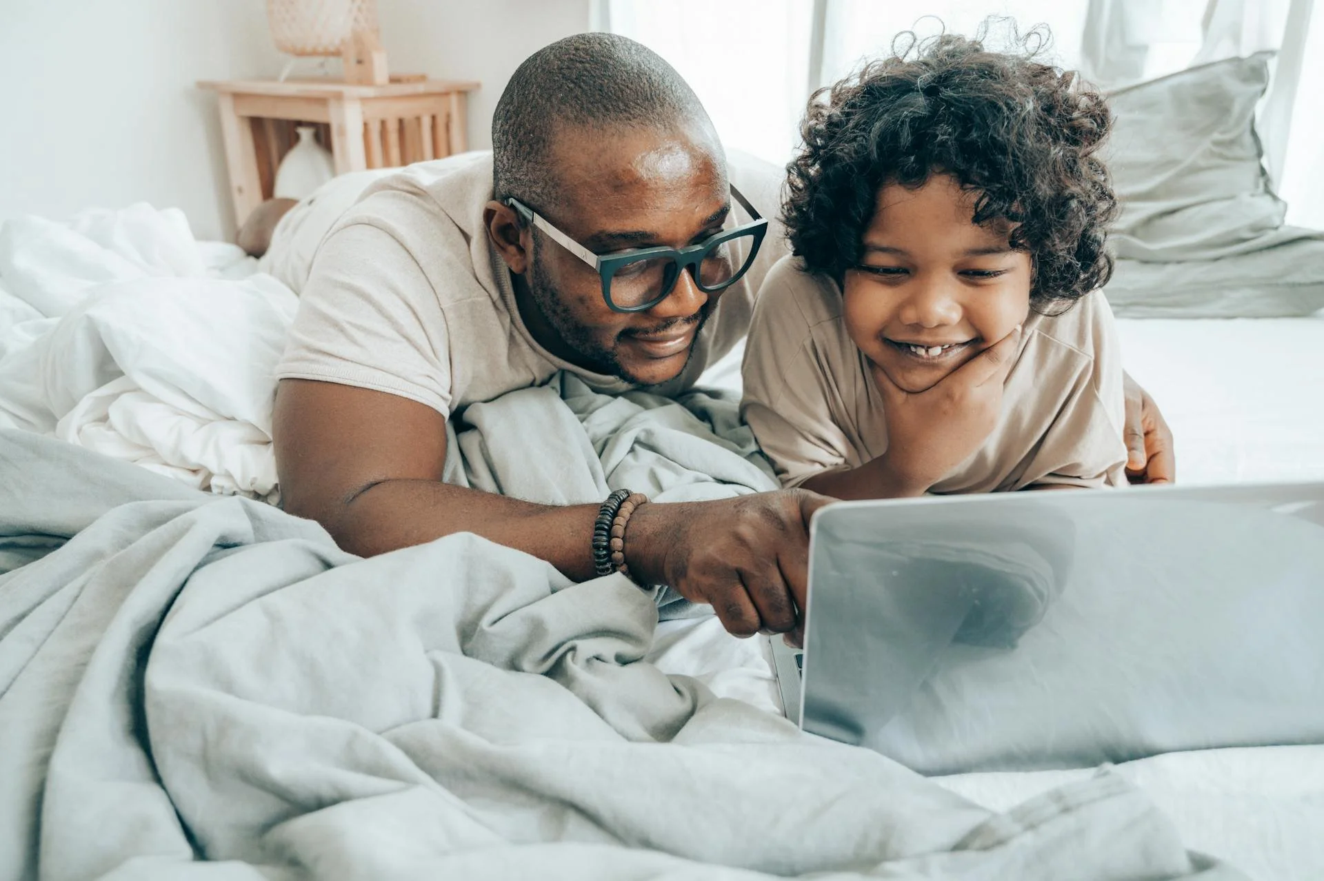 A man and boy lying on a bed, looking at a laptop screen, smiling and enjoying their time together.