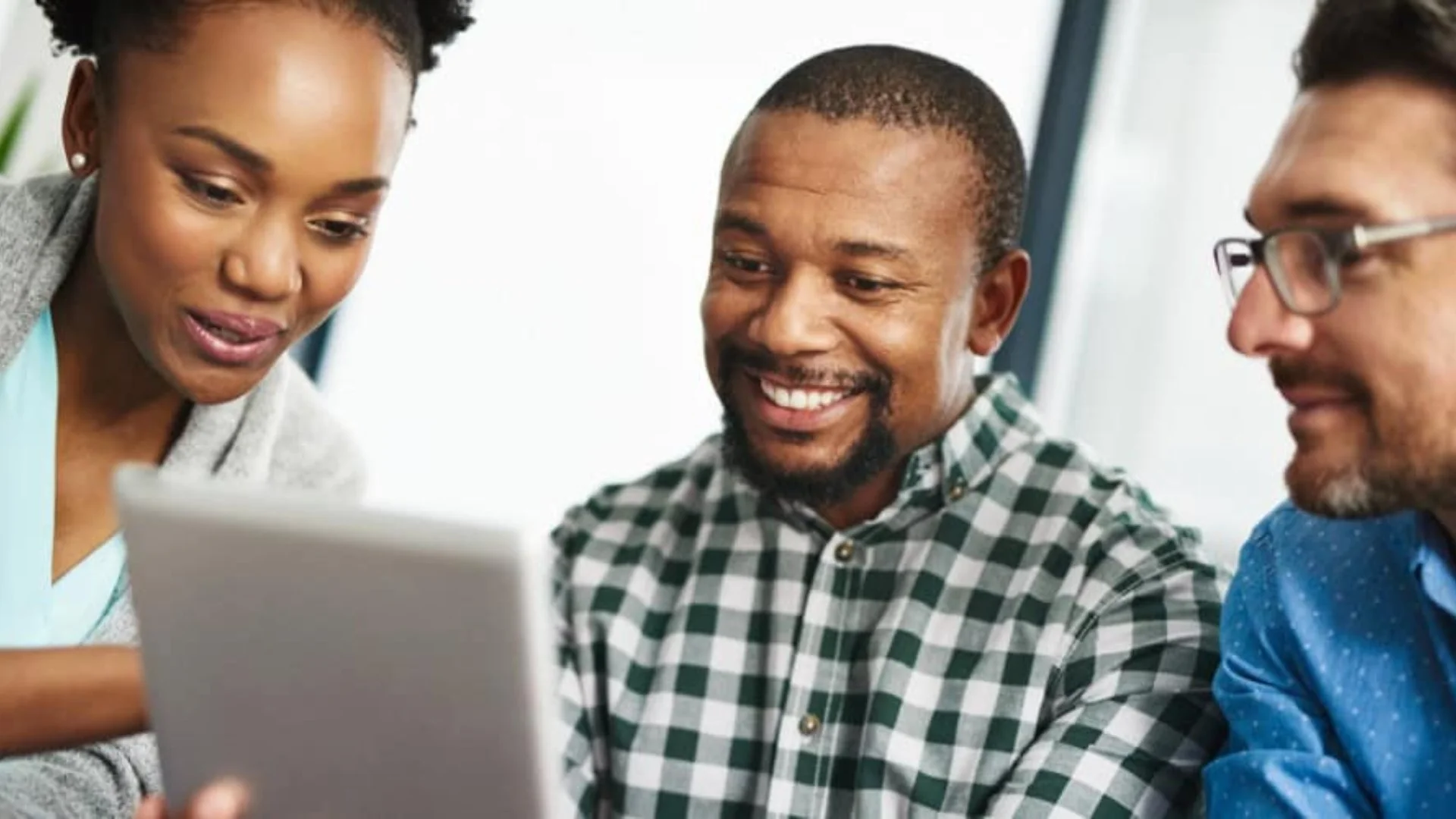 Three diverse people looking at a tablet, smiling, in a bright office setting.