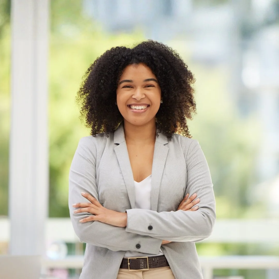A smiling woman with curly hair wearing a light gray blazer and crossing her arms, standing indoors with a blurred window and greenery in the background.