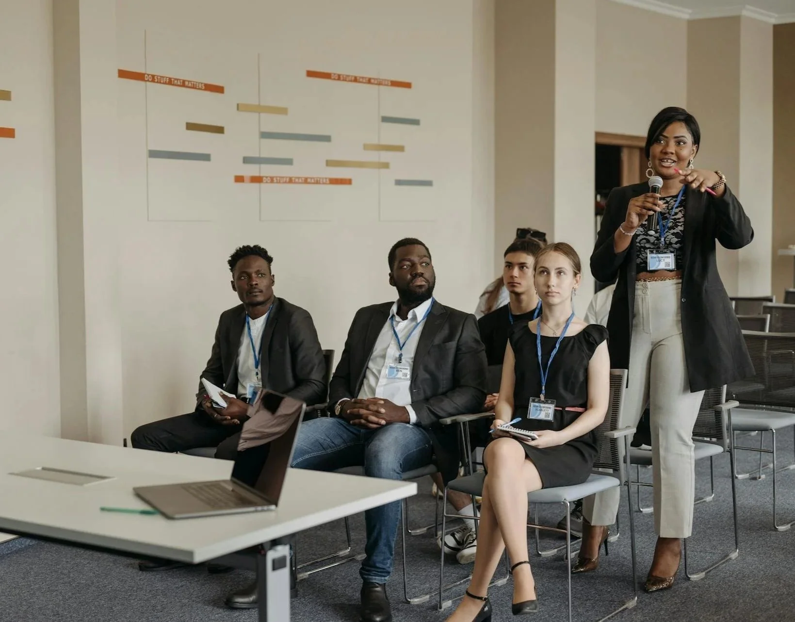 A woman wearing a black blazer and white pants is speaking into a microphone during a presentation or conference. She is gesturing with her hand and wearing a name badge. Four other people, two men and two women, are sitting and listening attentively in the background, also wearing name badges. The setting appears to be a modern conference room with a laptop on a table in the foreground and a wall with colored bars and text behind.