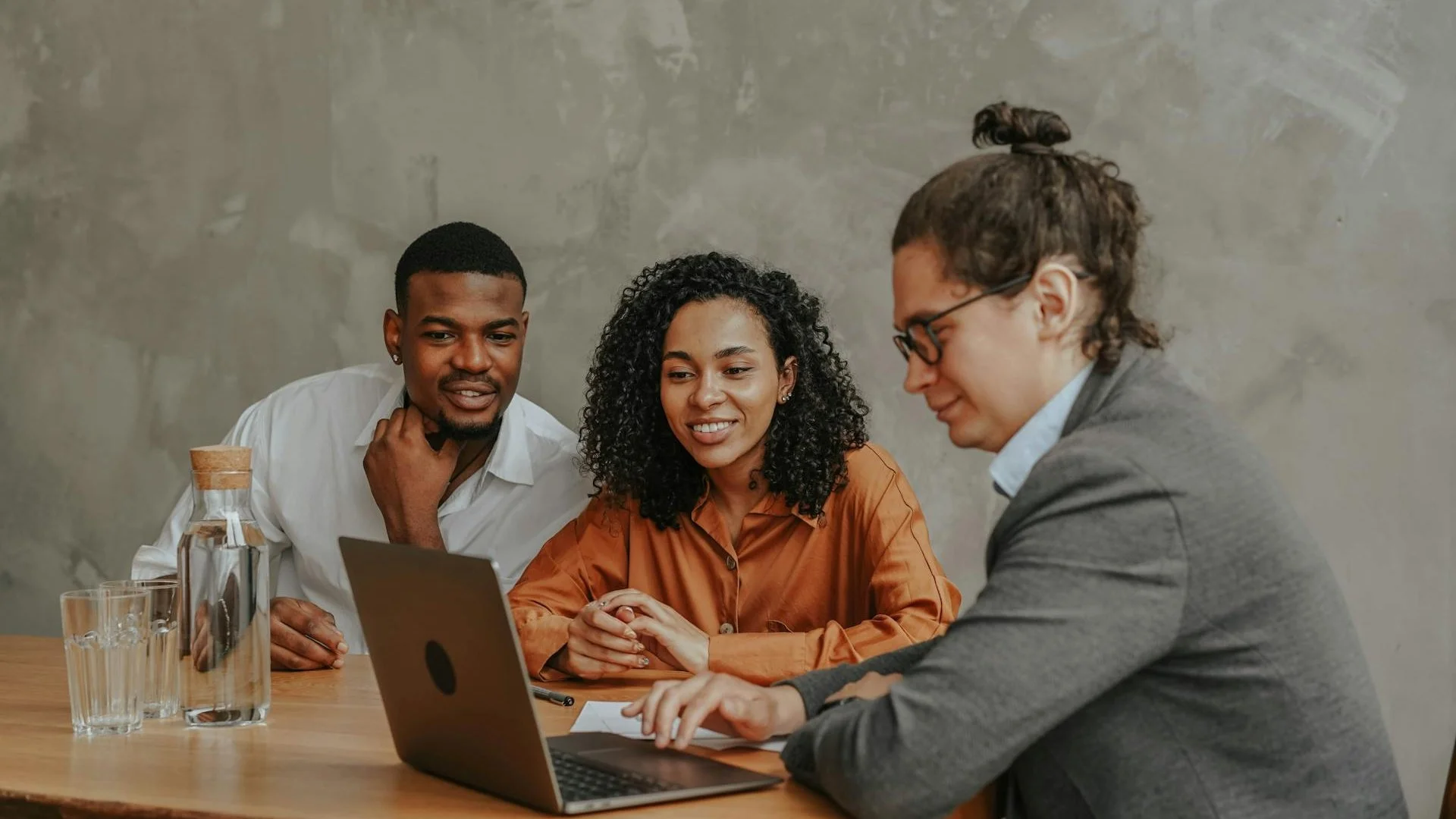 Three people sitting at a wooden table looking at a laptop, with a gray concrete wall in the background. Two glasses and a water decanter are on the table.