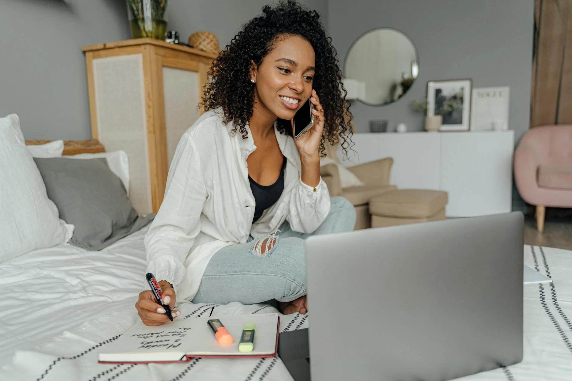 A young woman with curly hair, sitting cross-legged on a bed, talking on her cellphone, with a laptop in front of her, writing on a notebook, surrounded by colorful highlighters, in a cozy bedroom.