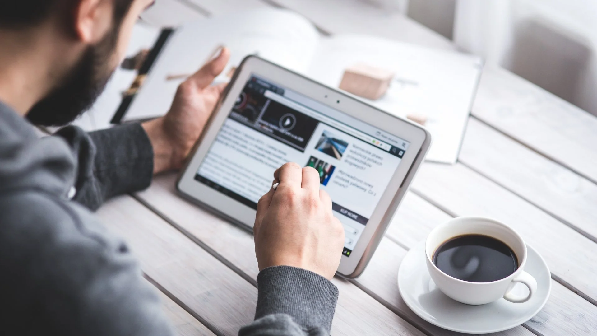 A man with a beard and gray jacket sitting at a white wooden table, holding a tablet and reading an article with a cup of black coffee nearby.