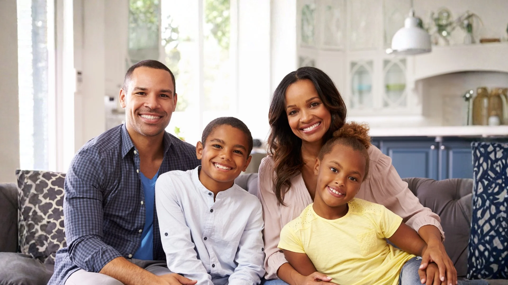 A happy multiracial family sitting close together on a sofa in a bright, modern kitchen, smiling at the camera.