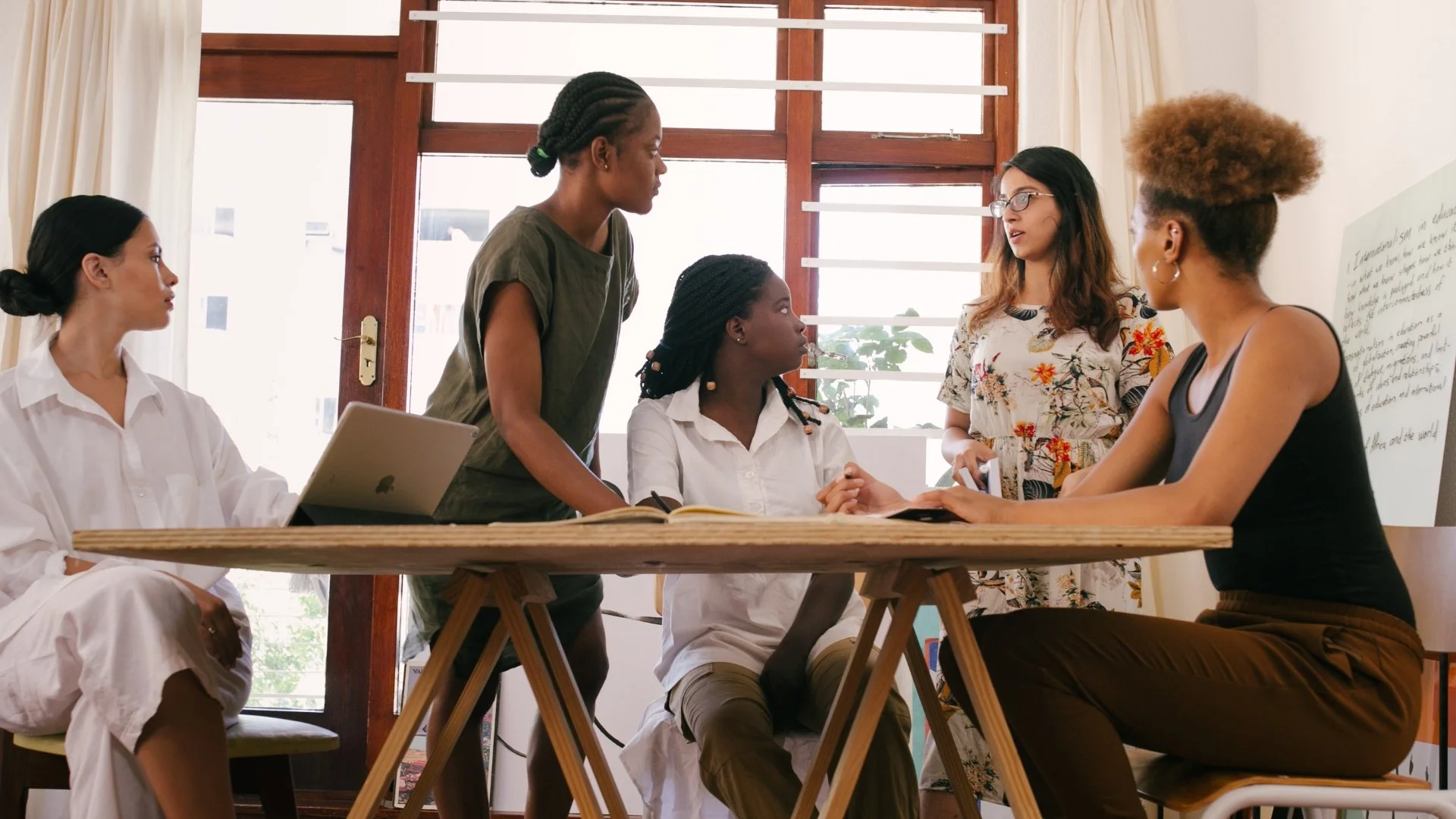 Six women in a discussion around a table in a room with large windows and wooden frames.