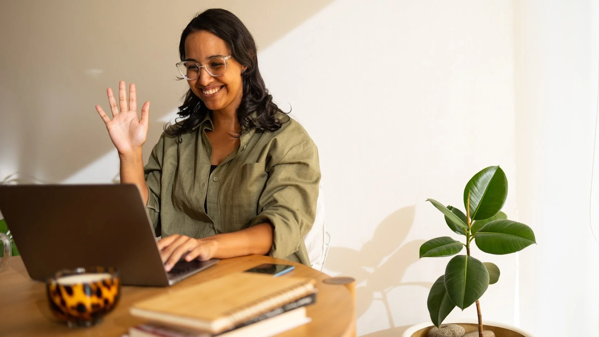 A woman with glasses and dark hair smiling and waving while sitting at a wooden desk with a laptop, a leopard print mug, a smartphone, and notebooks, next to a potted plant.