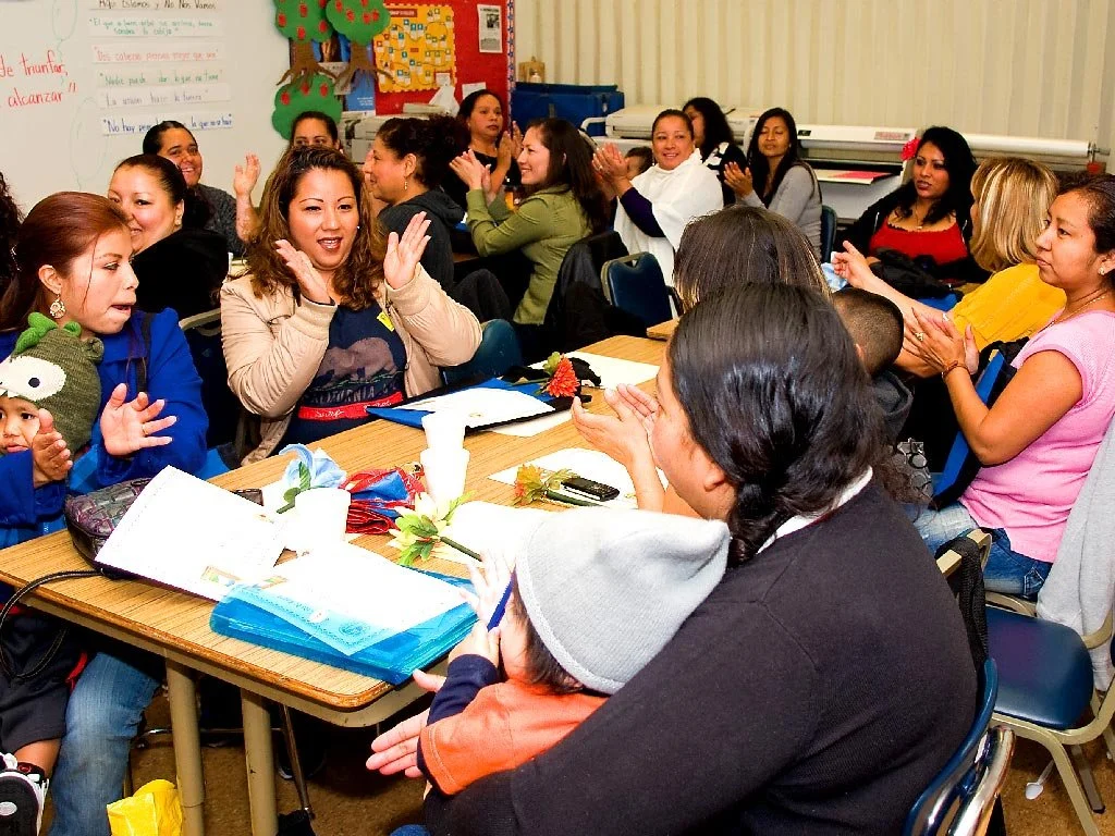 Group of women and children clapping and smiling around a table in a classroom or workshop setting.