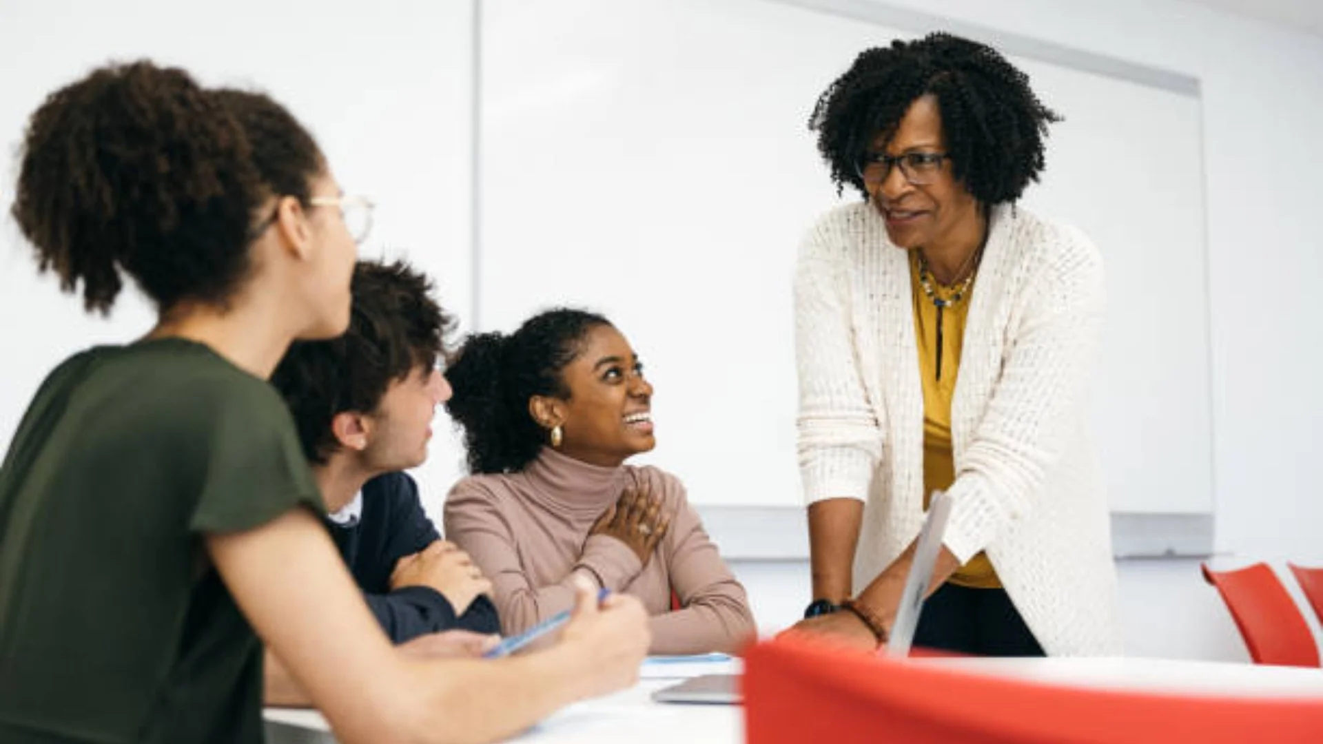 A woman standing and leaning over a table, speaking to four seated people in a meeting room.