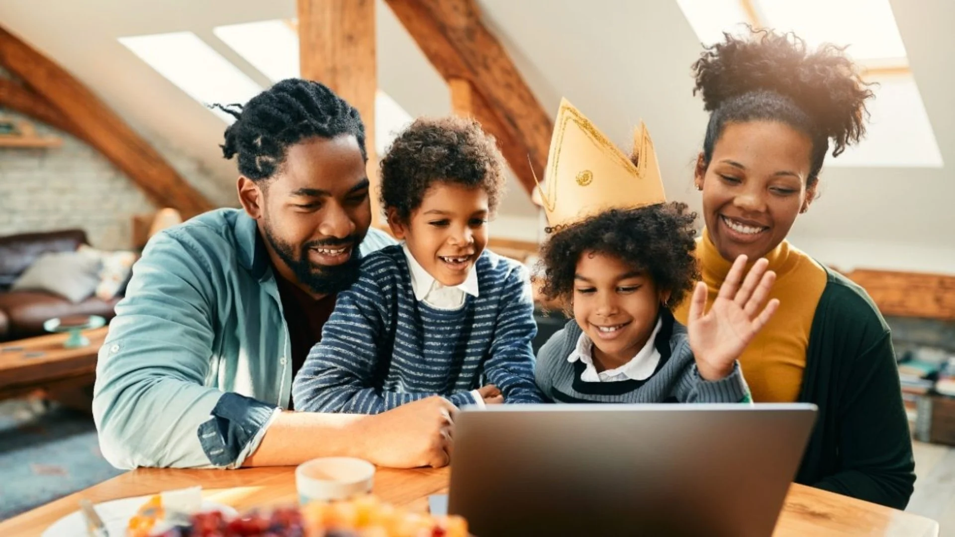 A multigenerational family of two adults and three children gathered around a laptop, smiling and waving at the screen, in a cozy attic space with wooden beams and natural light.