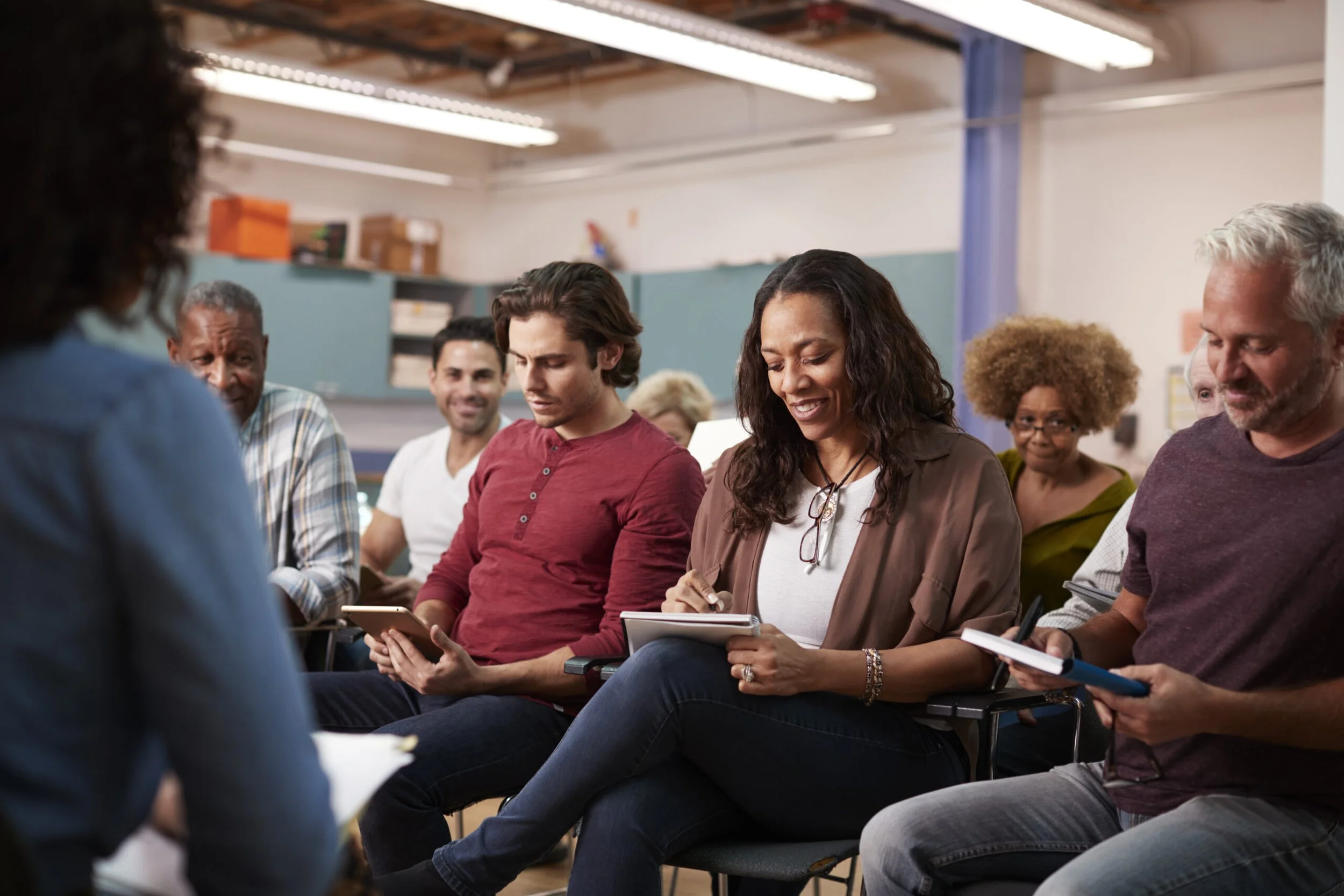 Group of diverse adults attending a workshop or meeting, seated in chairs, taking notes and listening attentively.