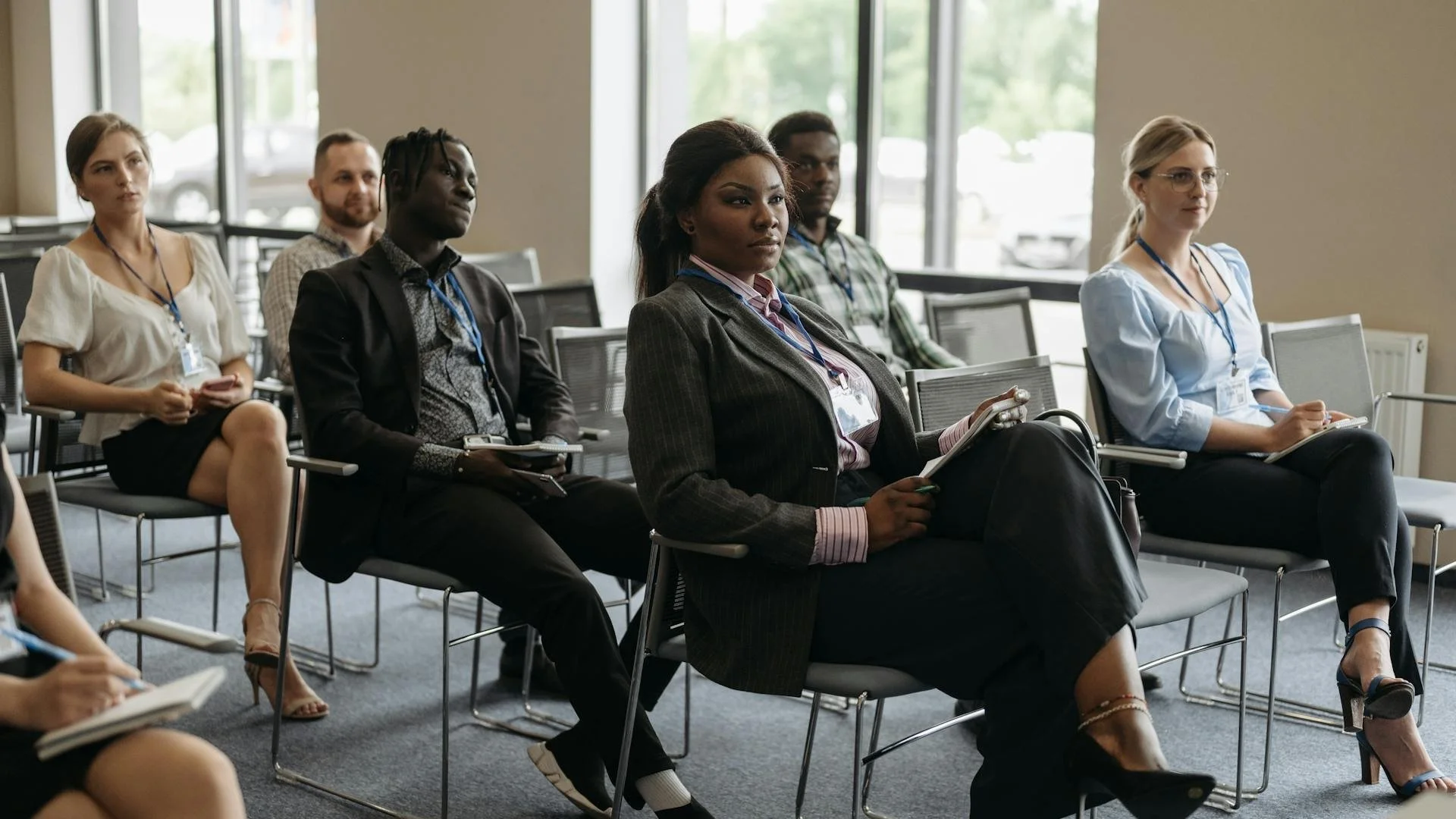A group of diverse people sitting in a conference room, attentive, some taking notes, with large windows in the background.