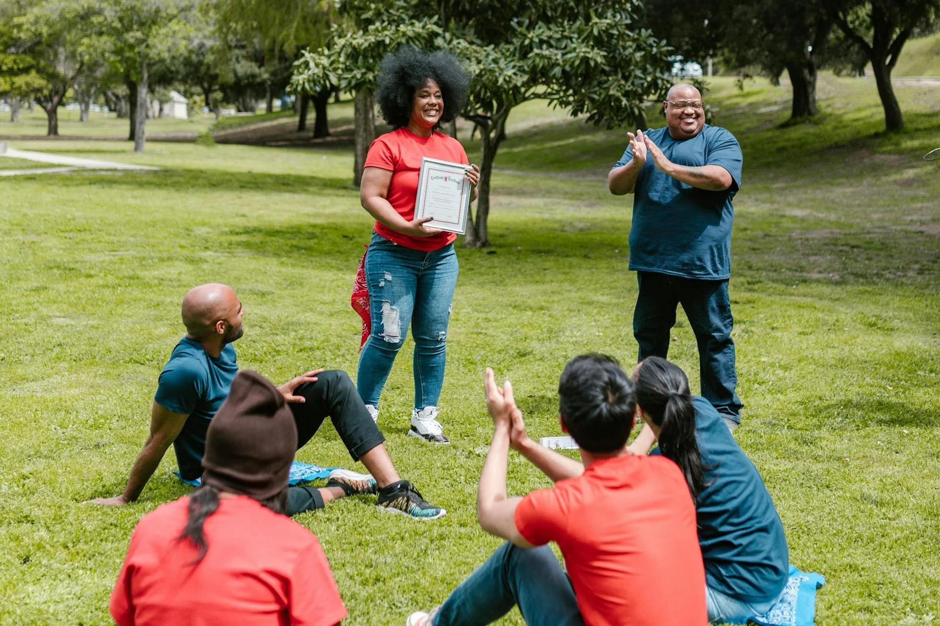 A woman holding a certificate smiles as she stands in a park, surrounded by people seated on the grass and clapping, with trees and greenery in the background.