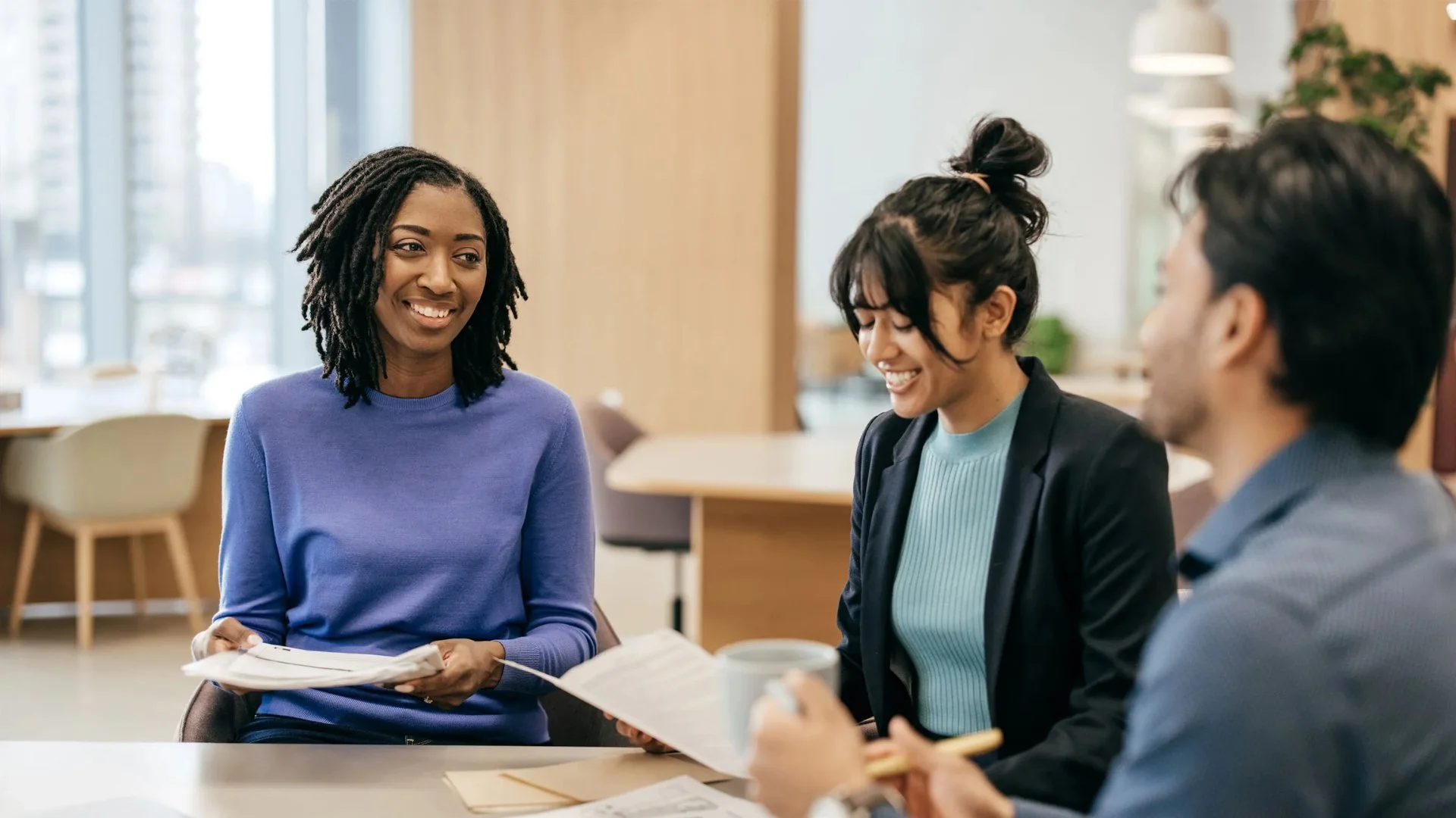 Three people in a meeting, two women and one man, all smiling and engaged in conversation, sitting at a table in a bright, modern office.