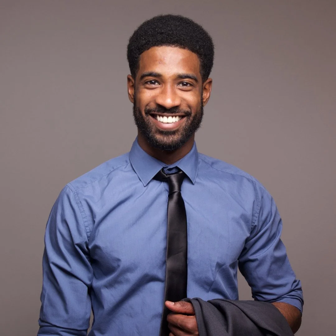 Smiling African American man wearing a light blue dress shirt and black tie, holding a suit jacket over his arm, against a gray background.