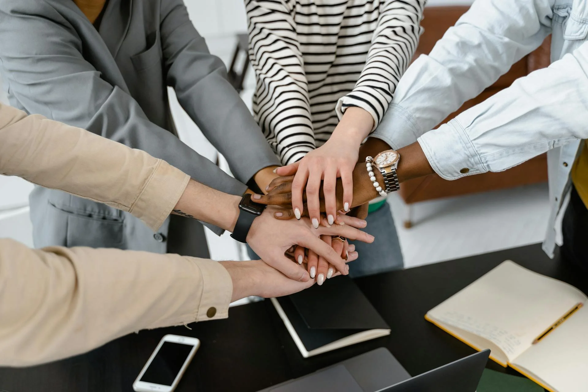 A group of diverse people stacking their hands together in teamwork over a dark desk with notebooks, a smartphone, and laptops.
