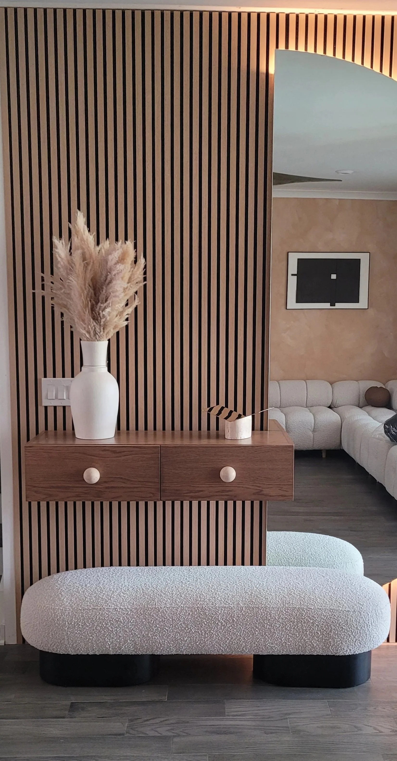 Interior view of a modern living space featuring a wooden slat partition, a wooden console with a white vase with pampas grass, and a patterned upholstered bench in the foreground. In the background, there's a beige sectional sofa and a wall with a black framed picture.
