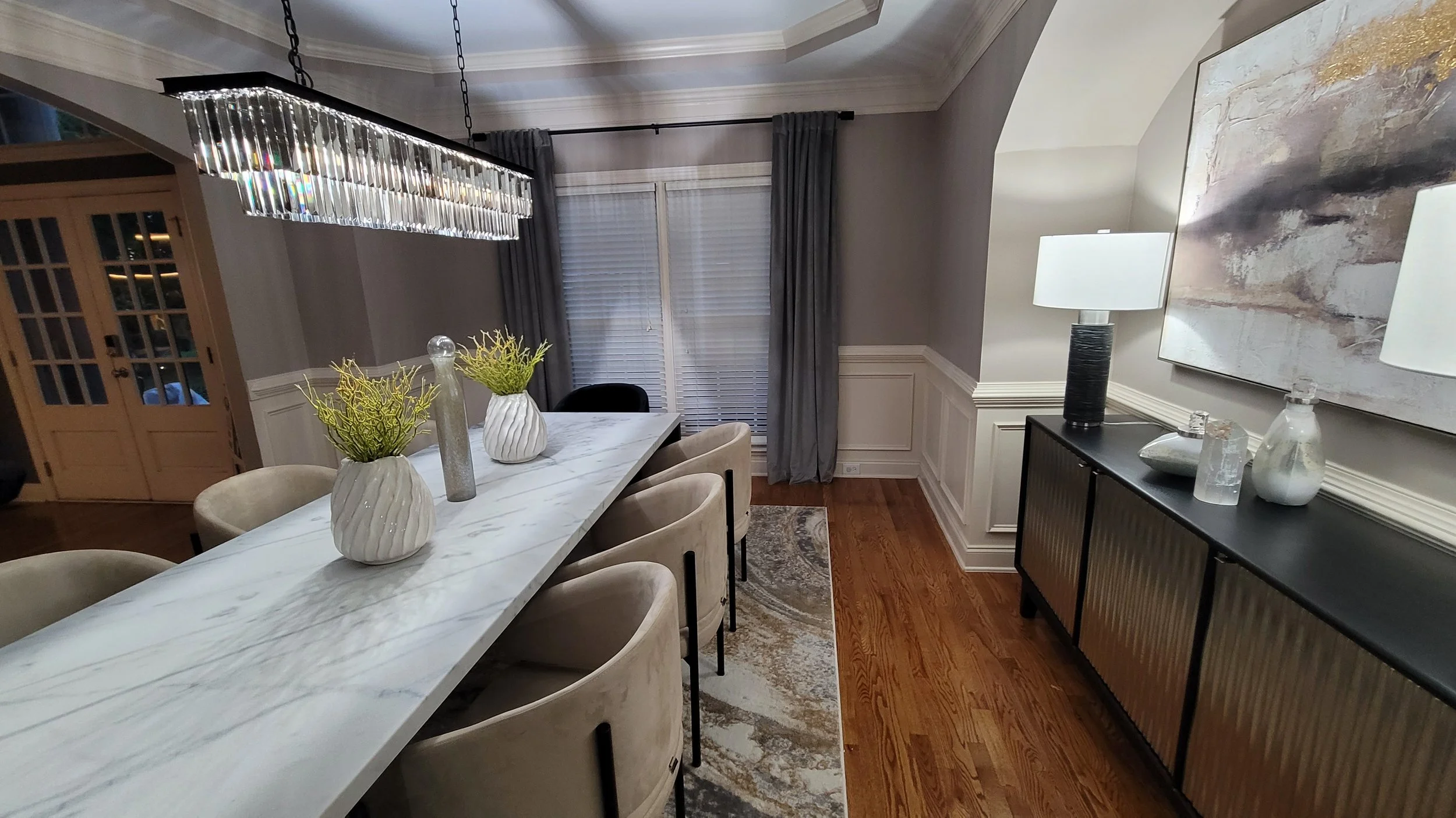 Modern dining room with a long white marble table, beige upholstered chairs, a glass chandelier, gray curtains, and a black sideboard with decorative vases and lamps.