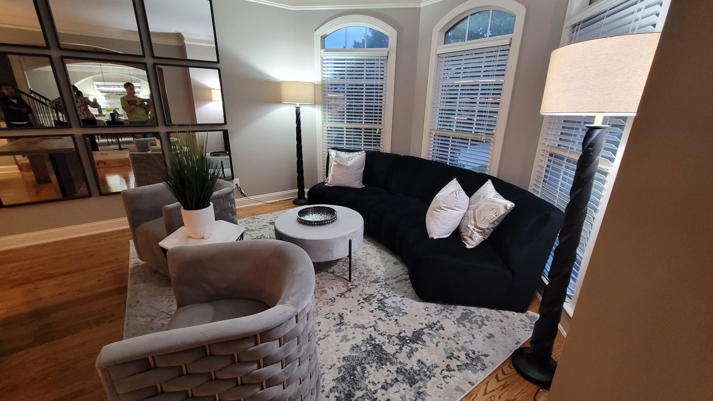 Living room with a black sofa, gray armchair, round white coffee table, tall black lamps, and large arched windows with white blinds. There is a rectangular mirror on the wall, a potted plant on the armchair, and a patterned rug on the wooden floor. People are visible in the reflection of the mirror.
