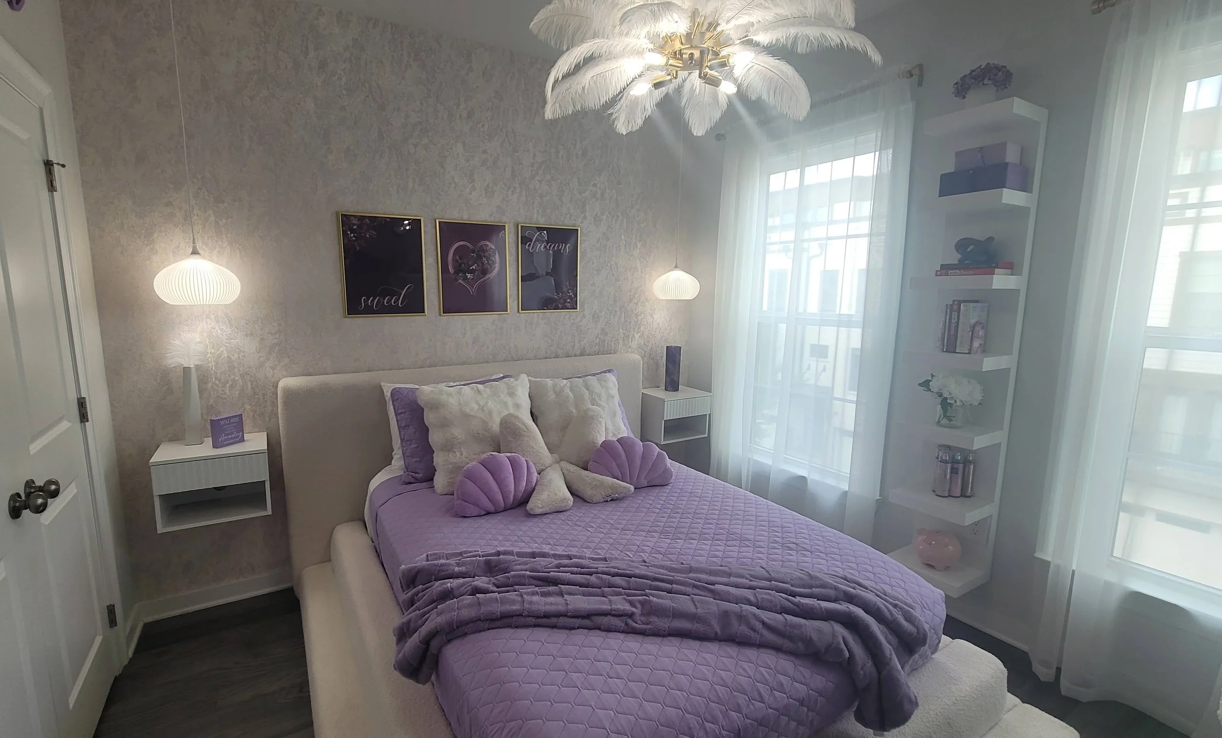 A cozy bedroom with purple bedding and plush pillows, artistic wall hangings, white side tables, and sheer curtains allowing natural light to brighten the space.