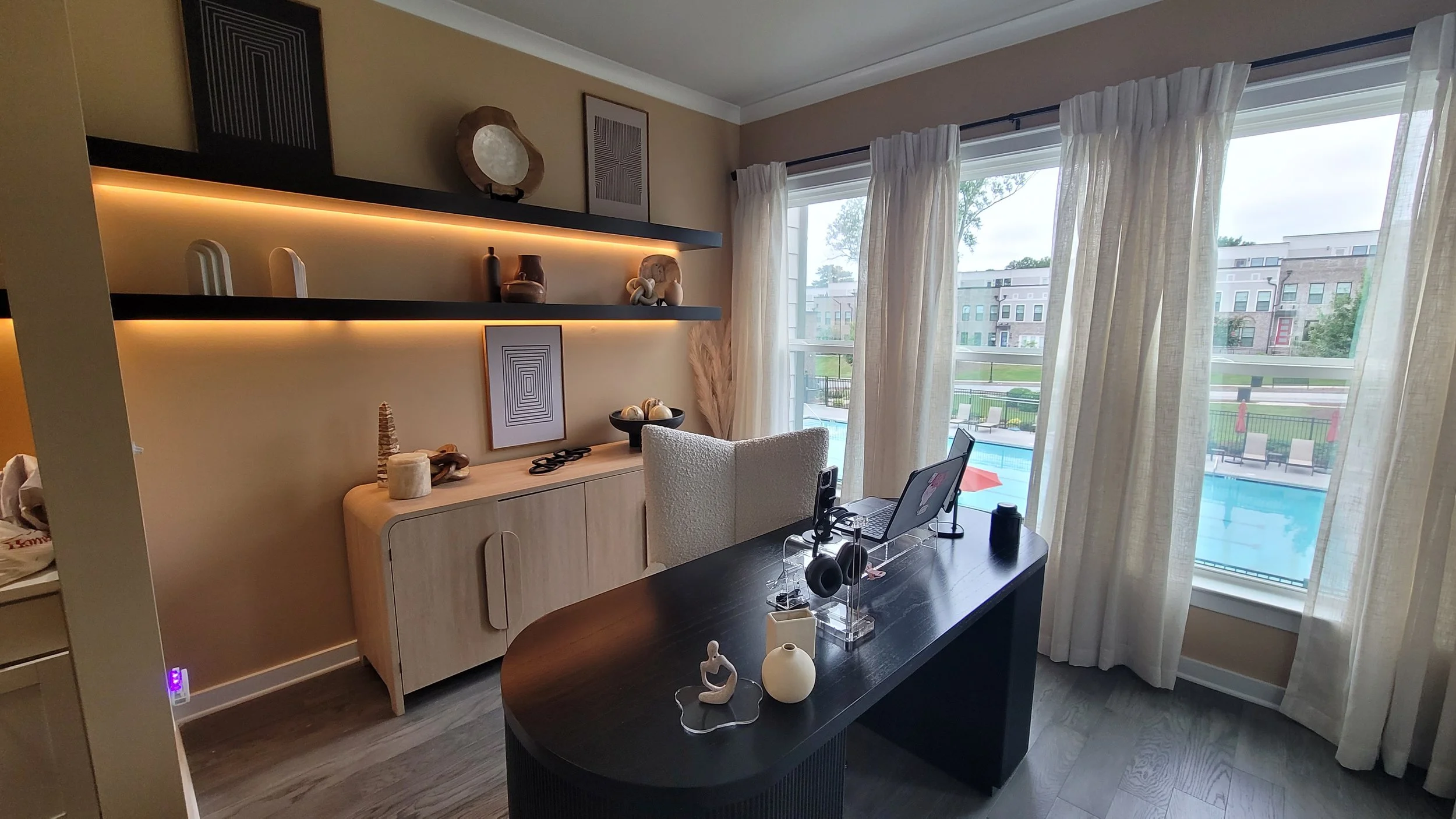 Modern home office with a black desk, white chair, decorative items, and large windows with sheer curtains overlooking a pool and community buildings.